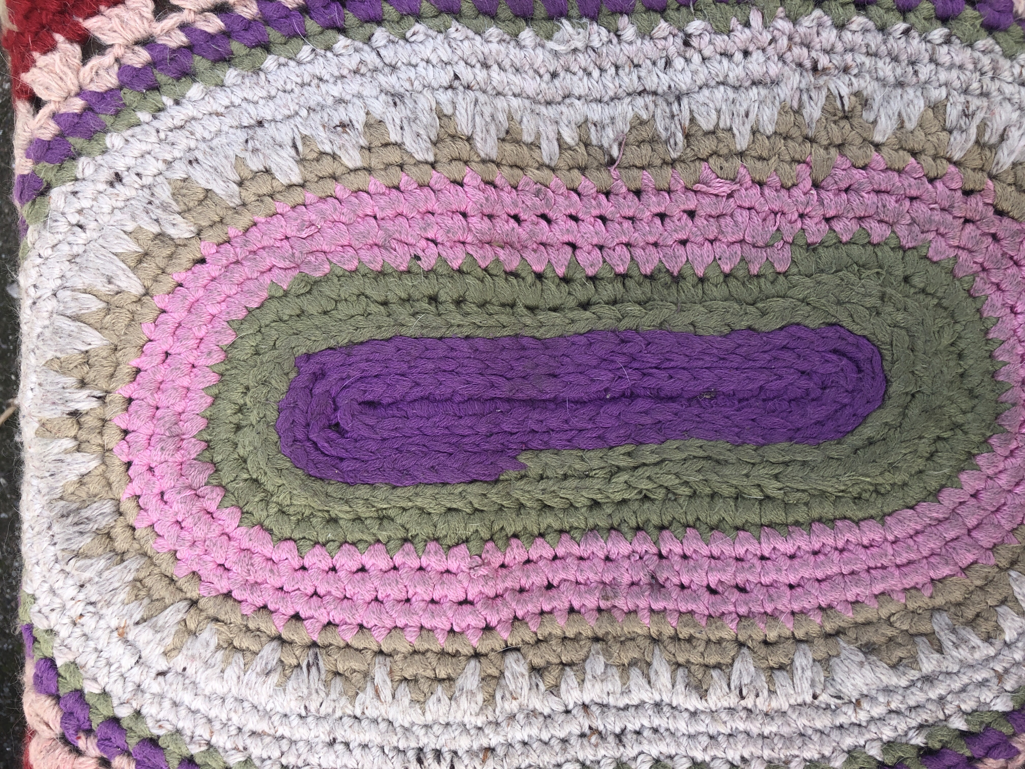 This is a close-up, overhead shot of a handmade, oval-shaped crochet rug. The rug is constructed from thick yarn in concentric oval bands. The innermost band is a vibrant purple, followed by an olive green band, then a pale pink band, and finally a light grey band. These bands form a radiating pattern, narrowing towards the center of the oval. The outermost visible sections of the rug show a mix of colors, including hints of maroon, purple, and lighter shades of pink and grey, interspersed with crochet stitching. The texture of the rug is visibly rough and tactile, indicating a rustic, handmade quality. The lighting suggests it is daytime, possibly outdoors given the slightly muted colors and the texture of the surface underneath. There are no people, text, or specific environmental cues to indicate a particular event or time of day beyond daylight. The location context provided, Пионерский, Russia, offers a general geographical setting but doesn't translate into any visual information within the image itself.