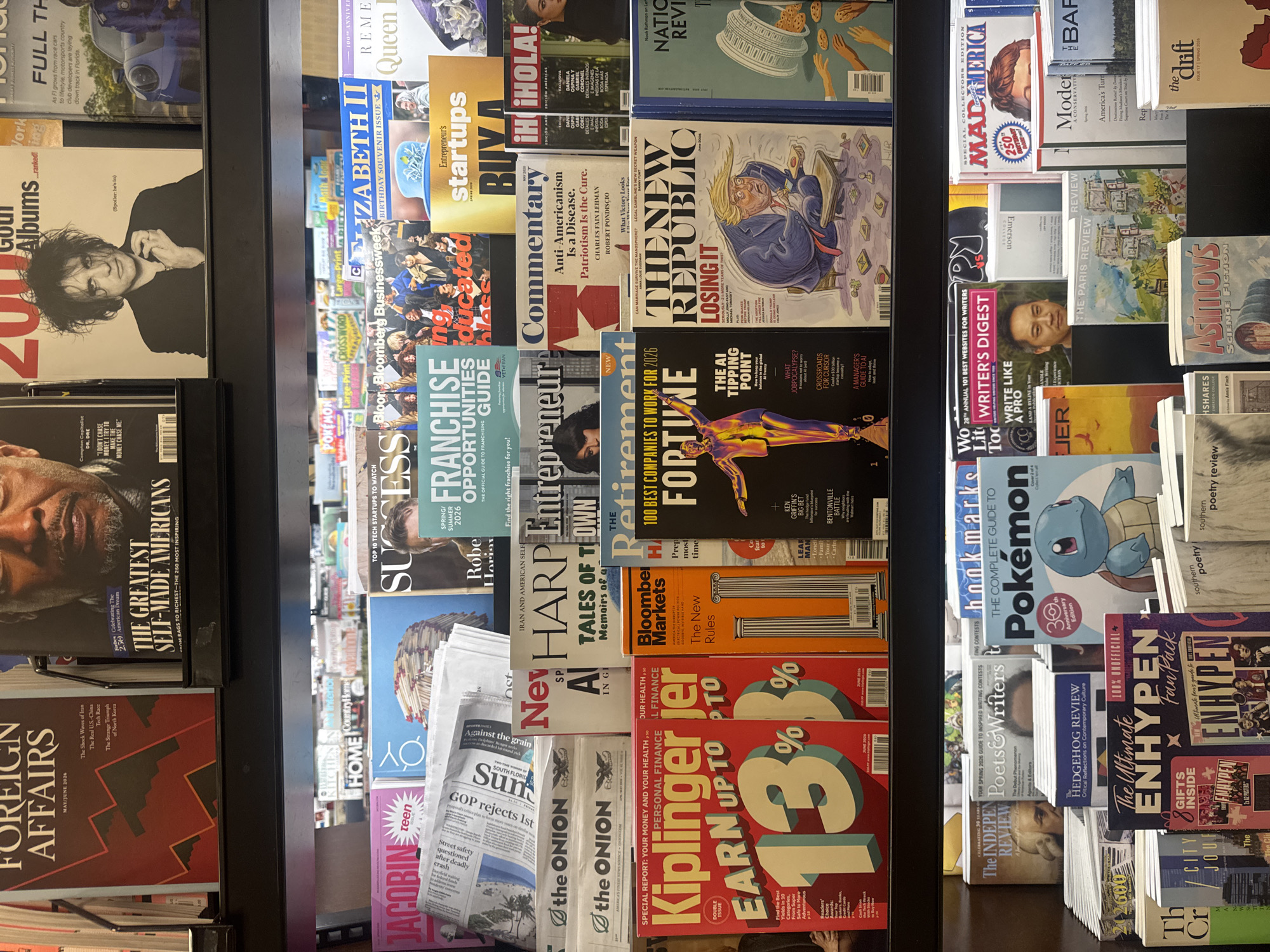 This image depicts a brightly lit, multi-tiered magazine rack inside a retail establishment in Boca Raton, United States of America. The rack is constructed of black metal shelving units, displaying numerous magazines and newspapers primarily oriented with their covers facing forward. No individuals are visible.
On the top shelf, from left to right, visible magazines include:
* *Foreign Affairs*, with a red and black abstract mountain design and "May/June 2024".
* A magazine titled *The Greatest Self-Made Americans*, featuring a close-up of a man's face.
* A magazine titled *200 Goth Albums ...ranked!*, featuring Robert Smith of The Cure on the cover, with "Spoiler: he's in".
* A partially obscured magazine titled *Full Thr...*, showing a blue sports car.
The second shelf from the top displays:
* *Jacobin Teen* with a pink cover.
* Several copies of *The Onion* newspaper.
* *Success Franchise Opportunities Guide* with a teal cover.
* *Entrepreneur Startups Buy A...* with a yellow cover.
* *Elizabeth II Birthday Souvenir Issue* with a white and gold design.
* *Remem... Queen D...*, partially obscured.
* Other partially visible titles like *Home*, *Country Home*, and *Bloomberg Businessweek*.
The third shelf from the top features:
* *Kiplinger's Personal Finance*, a double issue with a red cover promoting "Earn Up To 13%", dated June 2024.
* *Bloomberg Markets* with an orange cover featuring architectural columns.
* *Fortune 100 Best Companies to Work For 2026*, displaying a golden human-like figure and "The AI Tipping Point".
* *The New Republic Losing It*, dated June 2024, showing a cartoon illustration of Donald Trump.
* A partially visible *National Review* cover showing a cookie jar.
* Other magazines, including *New Harp*, *Commentary* ("Anti-Americanism is a Disease, Patriotism is the Cure."), *Hola!*, and *Retirement*.
The bottom shelf presents:
* *The Independent Review* with a black and white illustration.
* *Poets & Writers* showing a person's eyes.
* *The Hedgehog Review*.
* *Pokémon The Complete Guide*, featuring Squirtle and "30th Anniversary Edition".
* *The Ultimate ENHYPEN FanPack* with a purple cover and "8 Gifts Inside".
* *Writer's Digest* with a man's portrait on the cover.
* *The Paris Review* with an illustrated cover.
* *Asimov's Science Fiction*.
* *Mad America* labeled "250th Birthday Edition".
* Partially visible titles such as *Modern A Conservative*, *The Draft*, *City Journal*, and *Southern Poetry Review*.