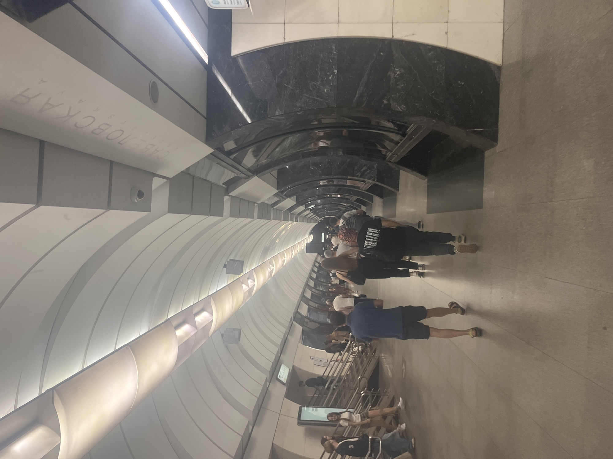 The image depicts the interior of a modern subway station in Moscow, Russia, likely Savelovskaya station, as indicated by the Cyrillic signage. The station features a high, arched ceiling with integrated lighting, creating a sense of spaciousness and illumination. The walls are a combination of white, curved panels and dark, reflective marble-like columns that curve inward, forming a series of arches.
A number of people are seen walking along the platform. They are dressed in casual attire, suggesting a normal day. In the foreground, a man in a blue polo shirt and dark shorts walks away from the camera, with a person in black further ahead also walking away. To the left, several individuals are walking towards the camera, including a woman in white shorts and a dark top, and a person in a blue jacket and jeans. Further down the platform, more people are visible, some walking in pairs or small groups.
The floor of the platform is paved with light-colored tiles. In the background, escalators and informational displays are visible. The overall atmosphere appears to be one of a busy but orderly public transportation hub.