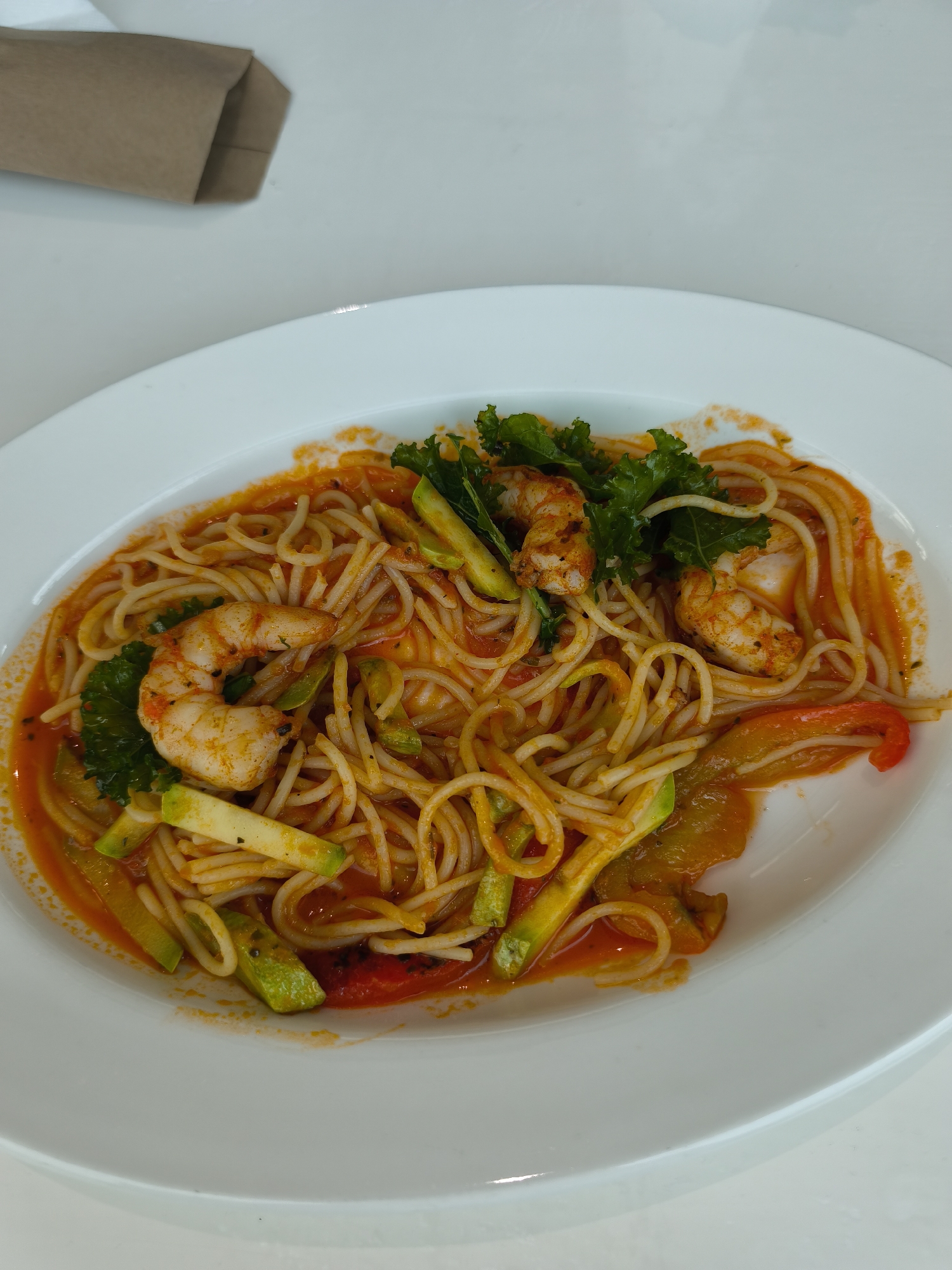 A close-up, full shot depicts a plate of shrimp pasta on a white table. The pasta appears to be spaghetti with a tomato-based sauce. Several cooked shrimp are visible among the pasta, along with slices of zucchini and red bell pepper. Some green leafy vegetables, possibly kale, garnish the dish. In the upper left corner of the frame, a brown paper bag is partially visible. The setting is nondescript, but the image’s data indicates a location within Moscow, Russia.