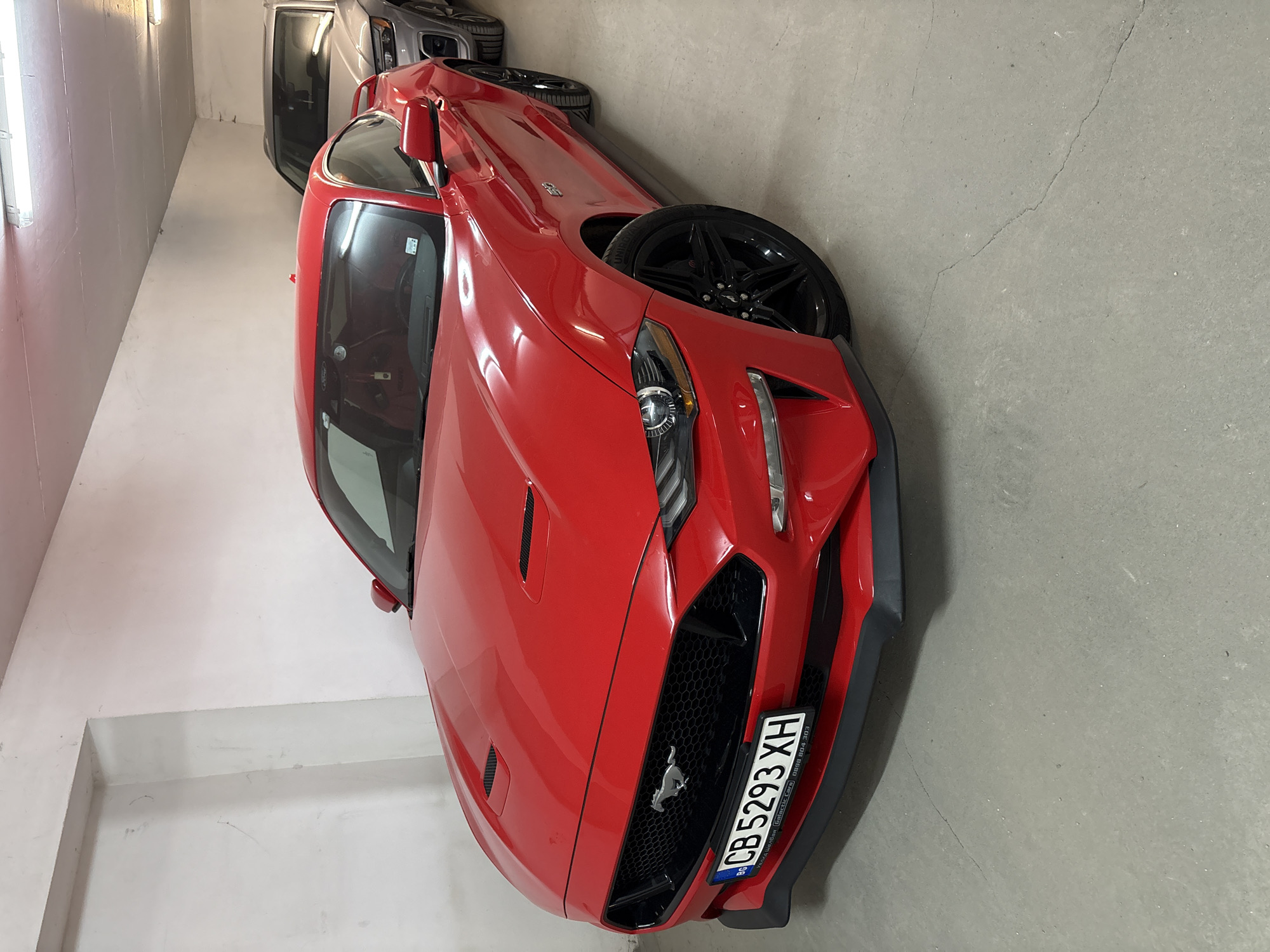 The image shows a red Ford Mustang parked in what appears to be an indoor parking garage or storage facility. The car's license plate reads "CB 5293 XH", which indicates it is registered in Bulgaria. The background includes portions of other vehicles, including a silver or gray SUV, suggesting a crowded storage situation. The parking area is mostly gray concrete. No people are visible in the image. It's impossible to confirm the exact location in Sofia, Bulgaria without further information or context.