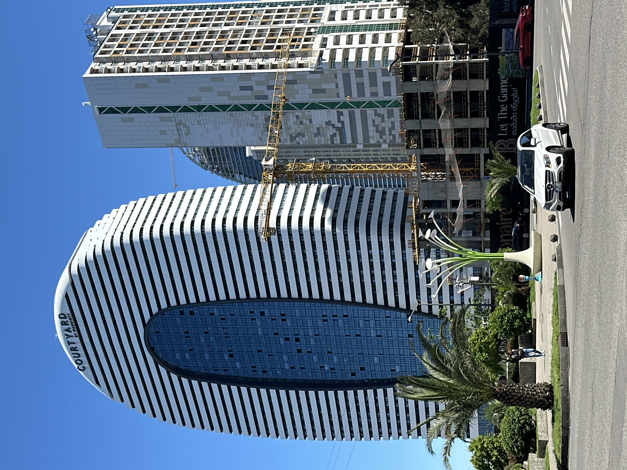 The image shows an architectural view in Batumi, Georgia, featuring a unique building with a horizontal oval void near the center, clad in white with dark vertical lines; the “COURTYARD BY MARRIOTT” brand is visible on the building. Another building, partially under construction with a yellow crane, stands in the background. The image is taken from street level, with palm trees and other greenery framing the lower portion, and what appears to be a modern art sculpture resembling stylized flowers is also present. A section of the road, with cars parked on the side, and a pedestrian walkway are also visible. Banners, "Let the Game (თამაში იწყება!)," on a billboard, can be seen. People can be seen walking along the sidewalk. The sky is clear and blue.