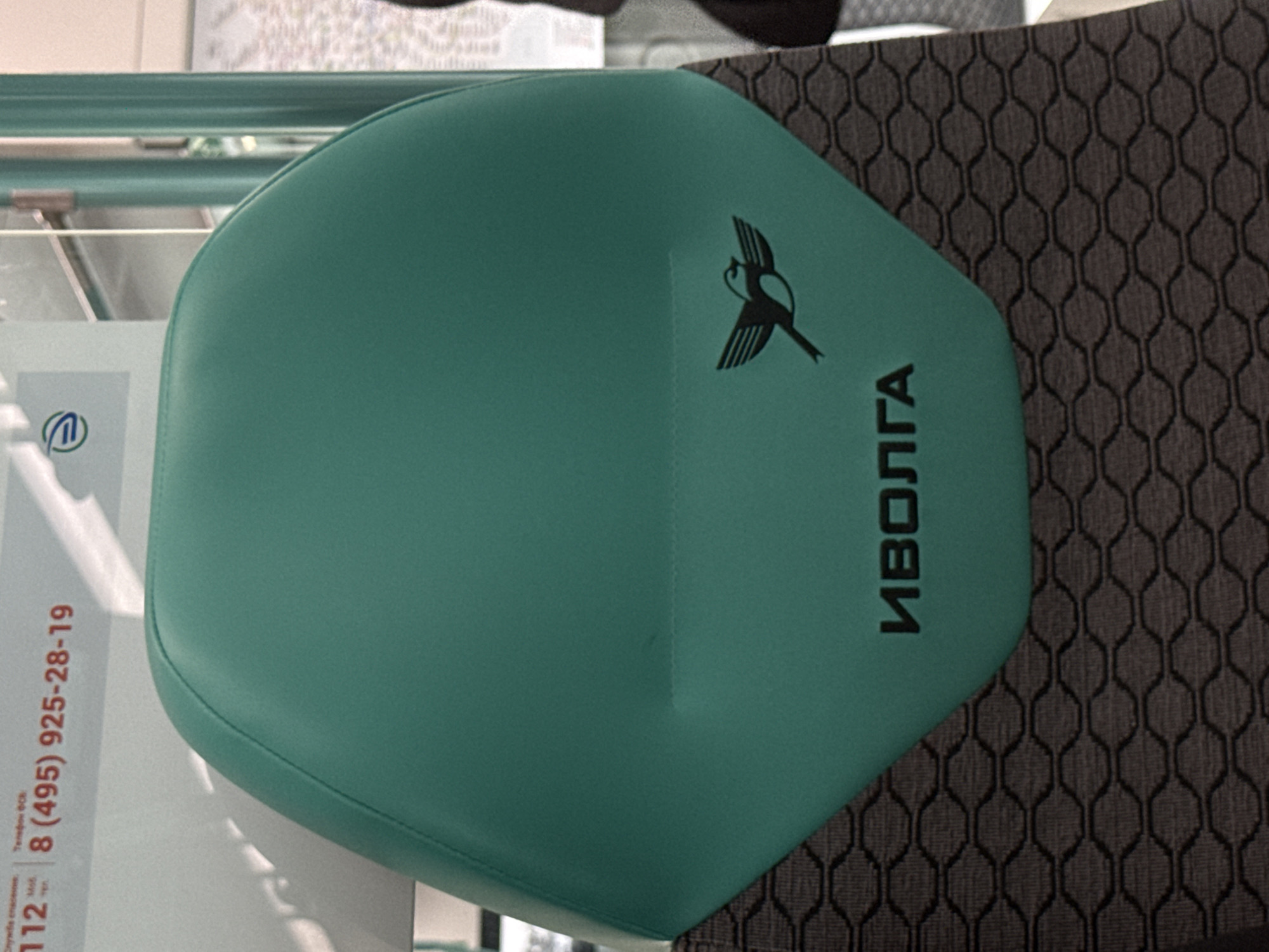 The image shows the back of a green seat on public transportation, likely a train or bus, in Moscow, Russia. The seat has a black logo of a bird in flight and the Russian word "ИВОЛГА" (IVOLGA), which translates to "Oriole" and is the name of a Russian electric train. In the background, a white panel with red lettering is visible, displaying emergency numbers "112" and a phone number "(495) 925-28-19". The seat is upholstered with a patterned dark fabric at the bottom. The lighting suggests an indoor environment with natural light filtering in, possibly from windows.