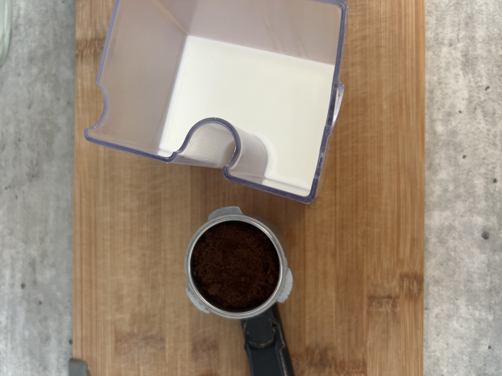A portafilter filled with dark brown coffee grounds sits to the left of a clear plastic container filled with a white liquid. Both objects are positioned on a light brown wooden cutting board, which is itself placed on a gray, textured surface. The scene appears to be indoors, with no discernible elements indicating a specific location in Volgograd, Russia. The lighting is even, suggesting overhead illumination.
