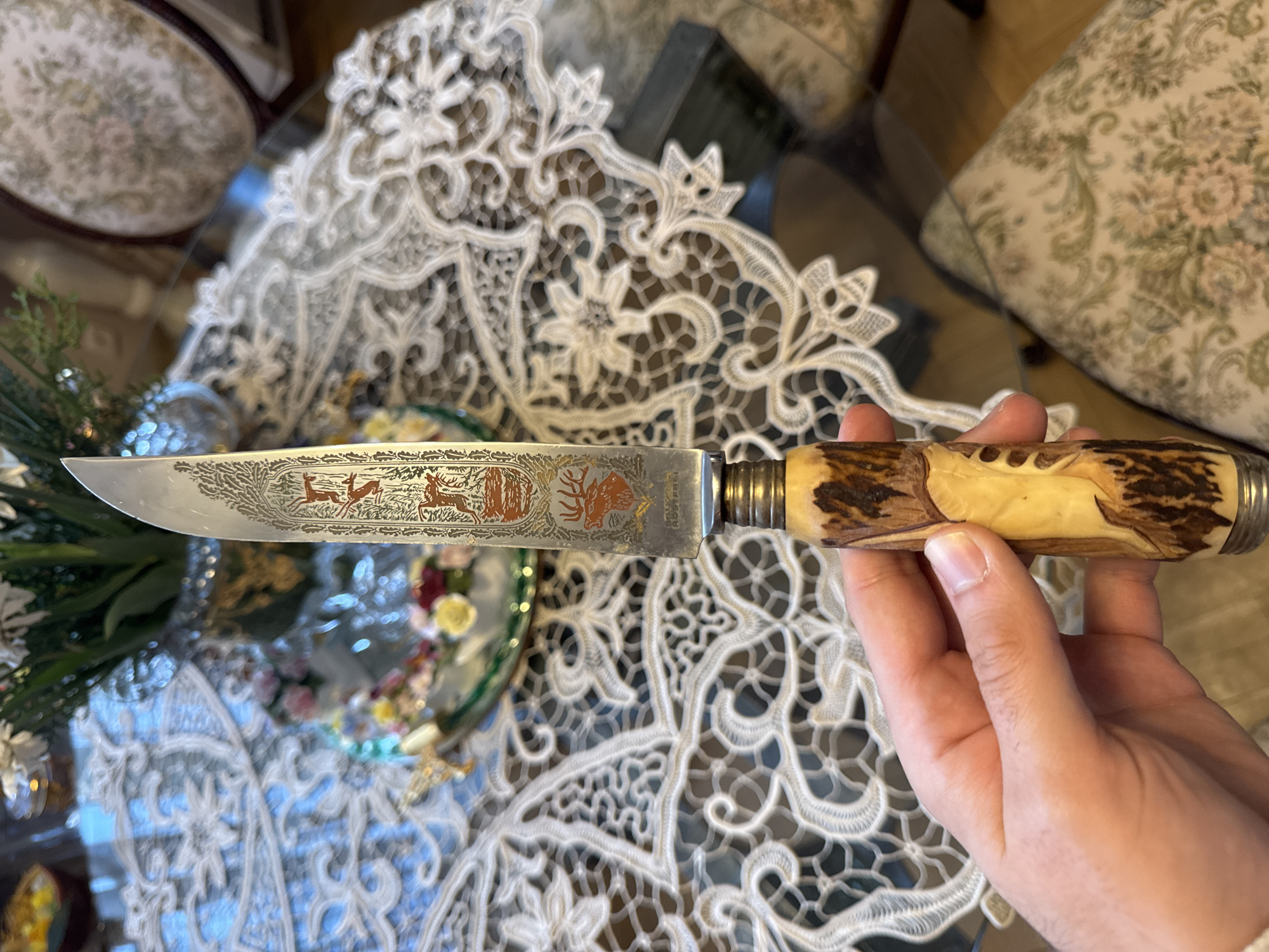 A person holds a knife with a stag antler handle and a blade etched with a forest scene including deer, against the background of a glass table covered with a white lace tablecloth. The table also has a small centerpiece containing flowers, greenery, and reflective glass elements. The location appears to be indoors, and furniture with floral upholstery is visible in the background. There is no immediate indication that the scene is specifically in Plovdiv, Bulgaria.