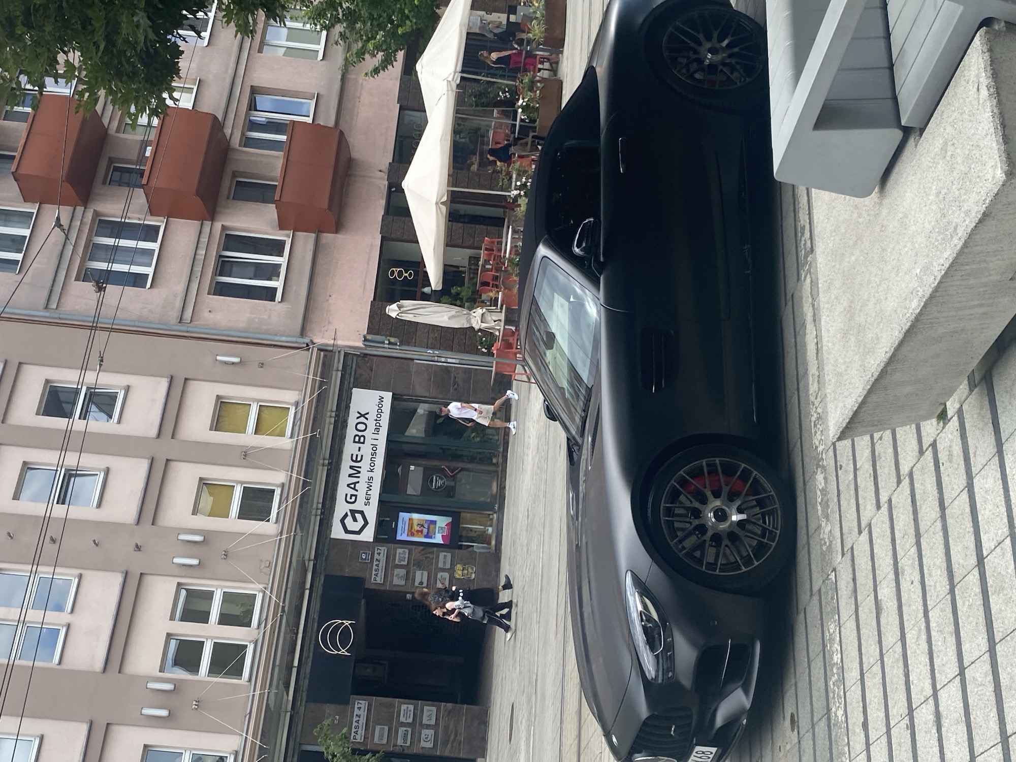 The image depicts a street scene in Poznan, Poland, with a matte black convertible sports car in the foreground. The car is parked on a cobblestone street with grey concrete benches in the immediate foreground. In the background, a multi-story building with a pinkish facade stands, featuring several windows and protruding red balconies. A commercial establishment with the name "GAME-BOX" is visible on the ground floor, advertising services for consoles and laptops. Several people are seen walking on the sidewalk in front of the building, and outdoor seating with white umbrellas is set up at a cafe or restaurant adjacent to the Game-Box store. The overall atmosphere suggests a daytime urban environment.