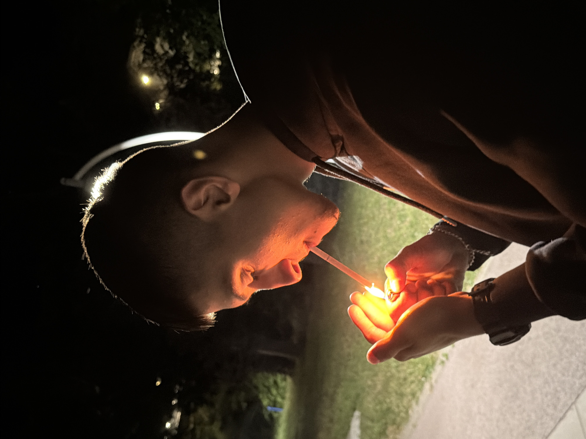 A young man is shown at night, close-up from the side profile, lighting a cigarette. He is wearing a dark hooded sweatshirt and a watch on his left wrist. The background is dark, with some blurred lights and greenery, suggesting an outdoor urban setting, possibly a park or street in Ruse, Bulgaria, illuminated by artificial lights. The flame of the lighter casts a warm glow on his hands and face.