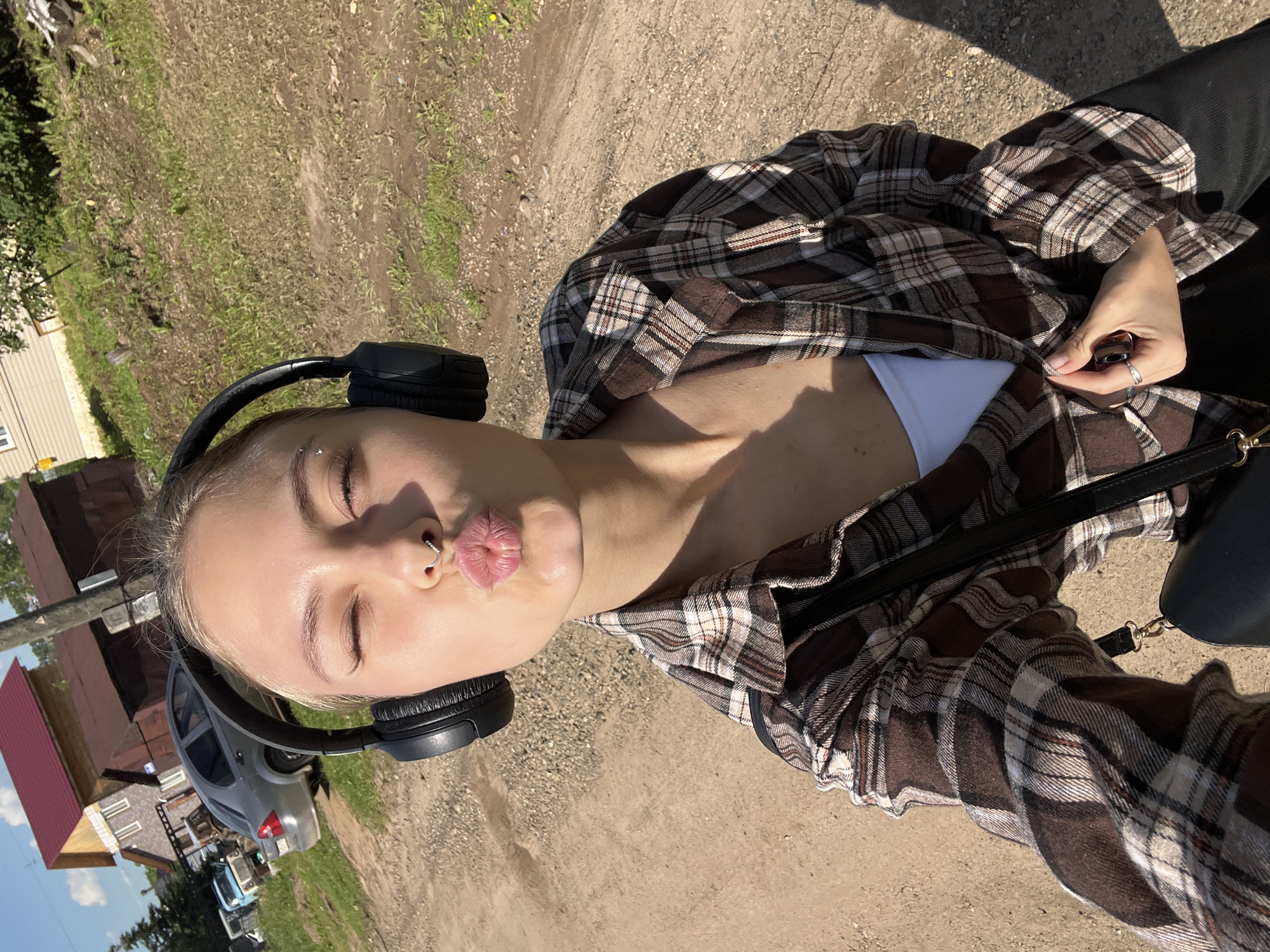 In the city of Foky, Russia, a young woman with blonde hair pulled back is taking a selfie outdoors. She is wearing large black headphones, a white tank top, and a brown and white plaid shirt. She has a septum piercing and her eyes are closed as she puckers her lips. She is wearing a ring on her finger. She is holding a black purse with a gold clasp. She is standing on a dirt path in front of a grassy embankment. In the background, there are several buildings and parked cars. The sky is blue with some clouds.