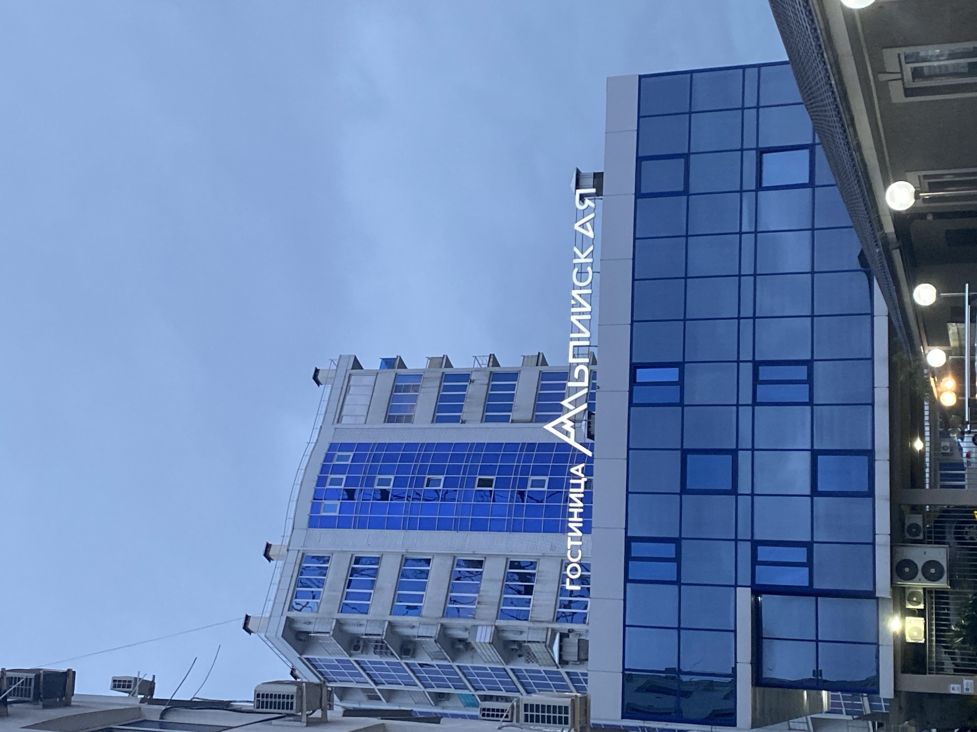 The image shows a building in Sochi, Russia. The building has a sign in Russian that reads "Гостиница Альпийская", which translates to "Alpine Hotel". The building is tall and has a modern design, with blue tinted windows and a white exterior. Part of an adjacent building is visible to the right, showing lights and air conditioning units. The sky is overcast.