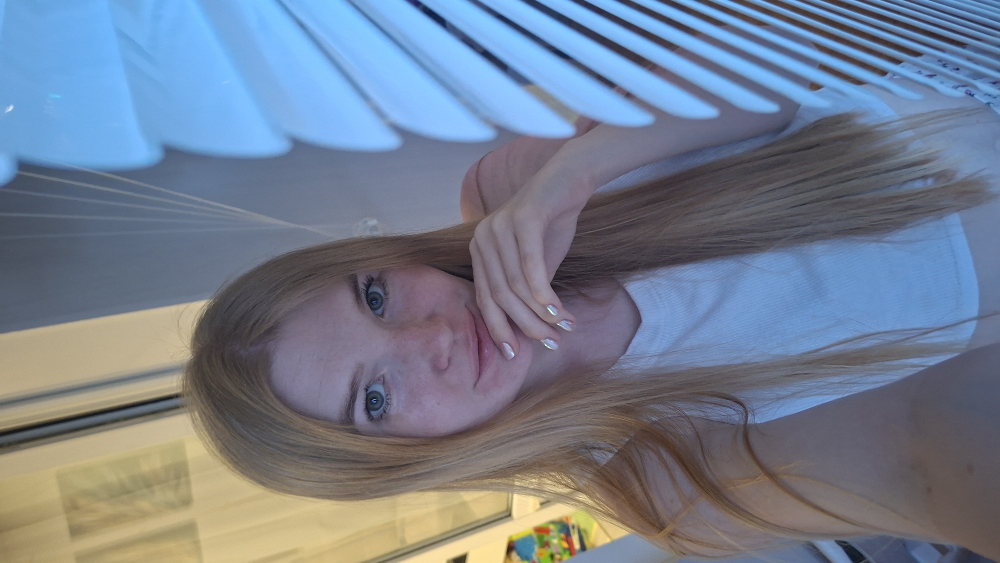 A fair-skinned person with light blonde hair and blue eyes is lying down. Their face is partially obscured by their hand. The person is wearing a white t-shirt. In the background, there are white blinds. The location may be a residence, as indicated by the presence of blinds and a colorful object that is potentially a child's toy. It is unclear if this is indeed the city of Chaikovsky, Russia.