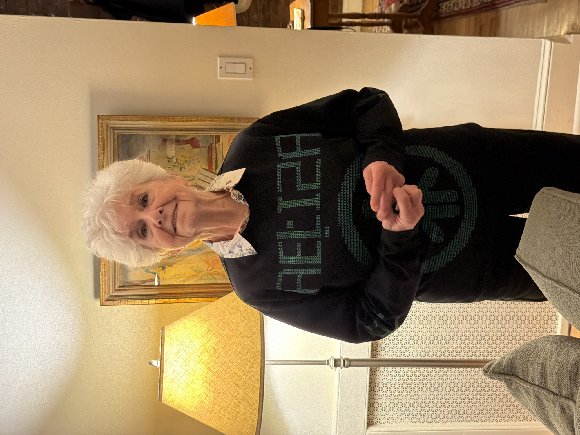 A mature woman with short white hair is indoors. She wears a black, long-sleeved top with green pixelated lettering, likely spelling a word or phrase, and a pattern or logo also constructed in green pixels. She is smiling slightly and has her hands clasped in front of her. Behind her is a wall adorned with a framed painting and a light switch. To the left of the woman, there is a lamp with a textured yellow shade. The lower right corner shows a portion of a gray couch. There is no direct evidence to identify the location as New York City or specify any interaction.