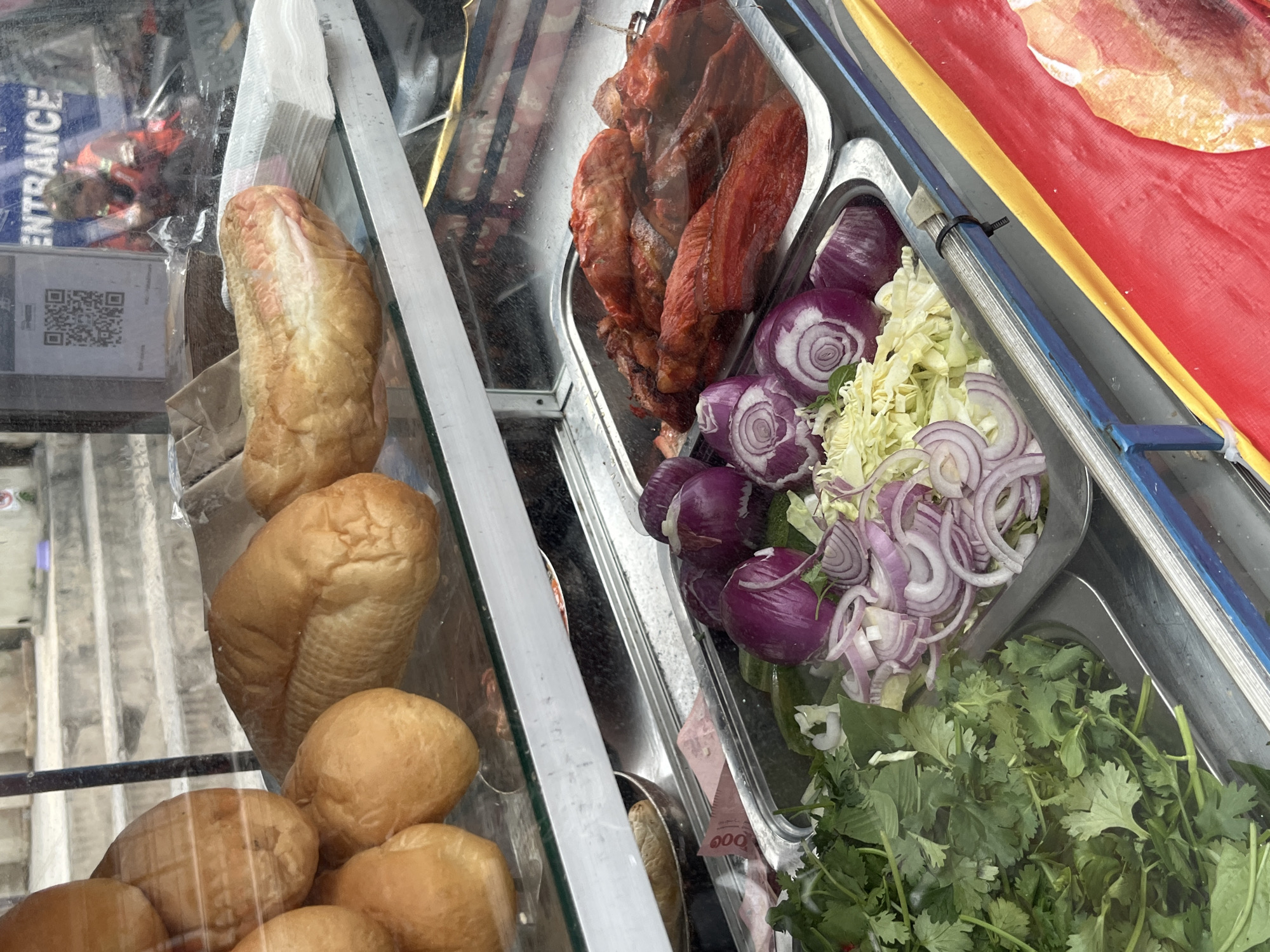 This image shows the contents of a display case likely used for selling food items, possibly for sandwiches or other similar dishes. The display case contains several types of bread, including baguette-style loaves and smaller, round buns. In addition to bread, the case holds various fillings and toppings. There is a tray containing roasted meat, sliced red onions, shredded cabbage, and fresh cilantro. The items are neatly arranged in their respective trays within the display case. The case is positioned outdoors, possibly on a street in Pattaya City, Thailand. The backdrop shows blurry images of people and buildings.
