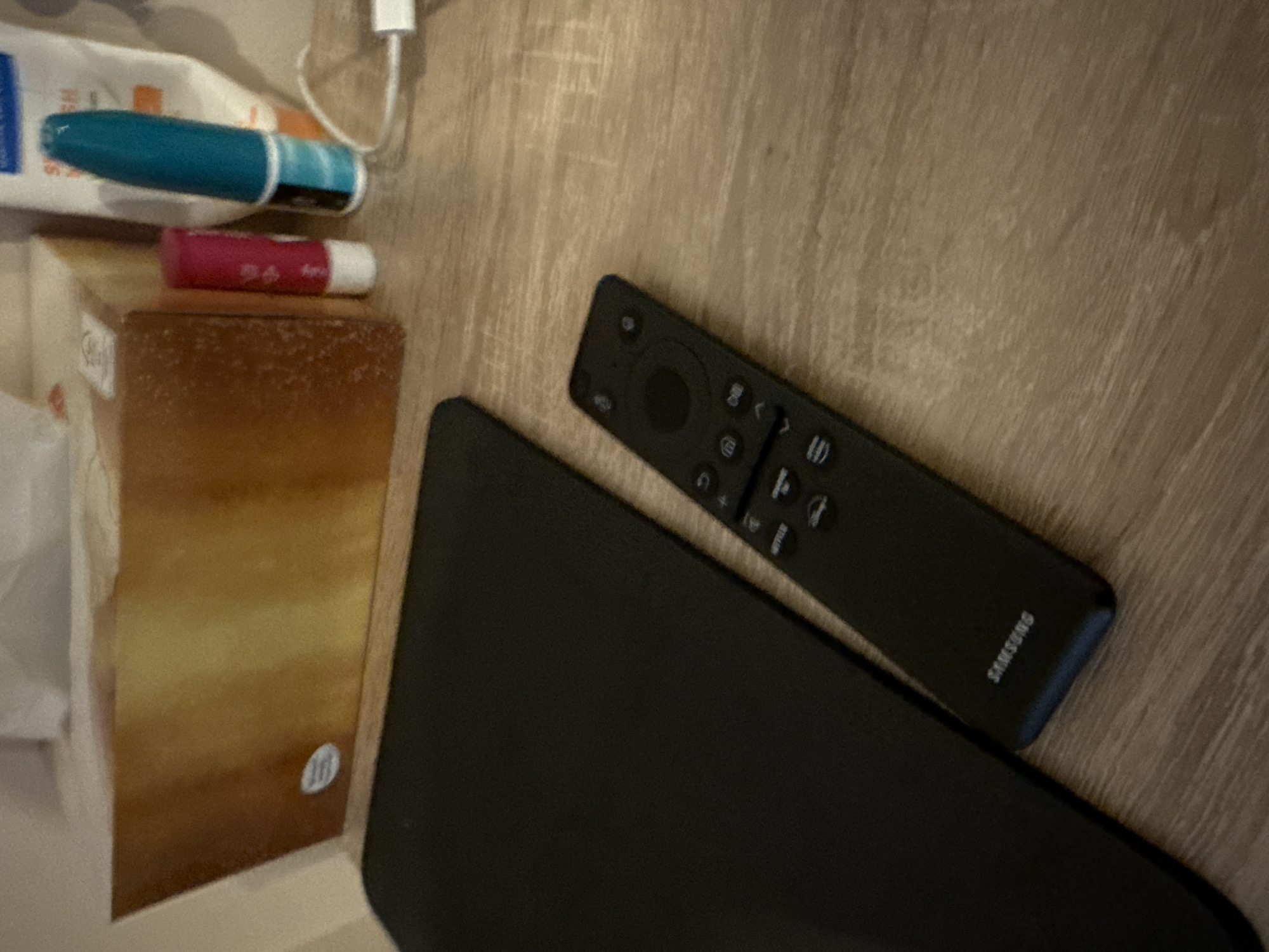 The image shows a tabletop scene. A black laptop partially covers a black Samsung TV remote. Beside them is a tissue box with a small pink and white lip balm and a blue pen on top. A white charger cable is visible near the tissue box. The surface appears to be a wooden table. There is no information to establish that the image was taken in Lovech, Bulgaria.