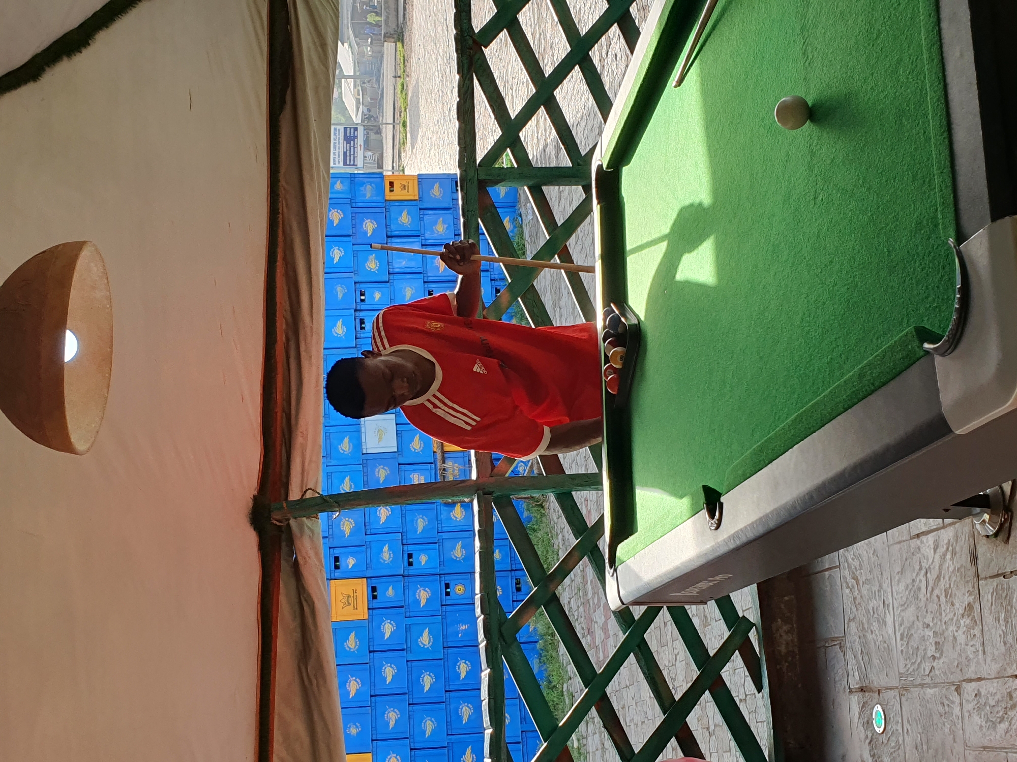 A young man in a red Manchester United jersey stands over a green pool table, holding a pool cue. He is positioned to the right of the frame, looking down at the pool balls arranged in a rack on the table. The pool table is situated outdoors under a thatched roof structure. Behind the pool table, a wall of blue plastic crates is visible, stacked high and displaying yellow logos. A green wooden railing separates the pool table area from the background. In the distance, beyond the crates and railing, a street scene with buildings and people can be observed. The overall lighting suggests it is daytime, with sunlight casting shadows. The location is identified as Lokoja, Nigeria.