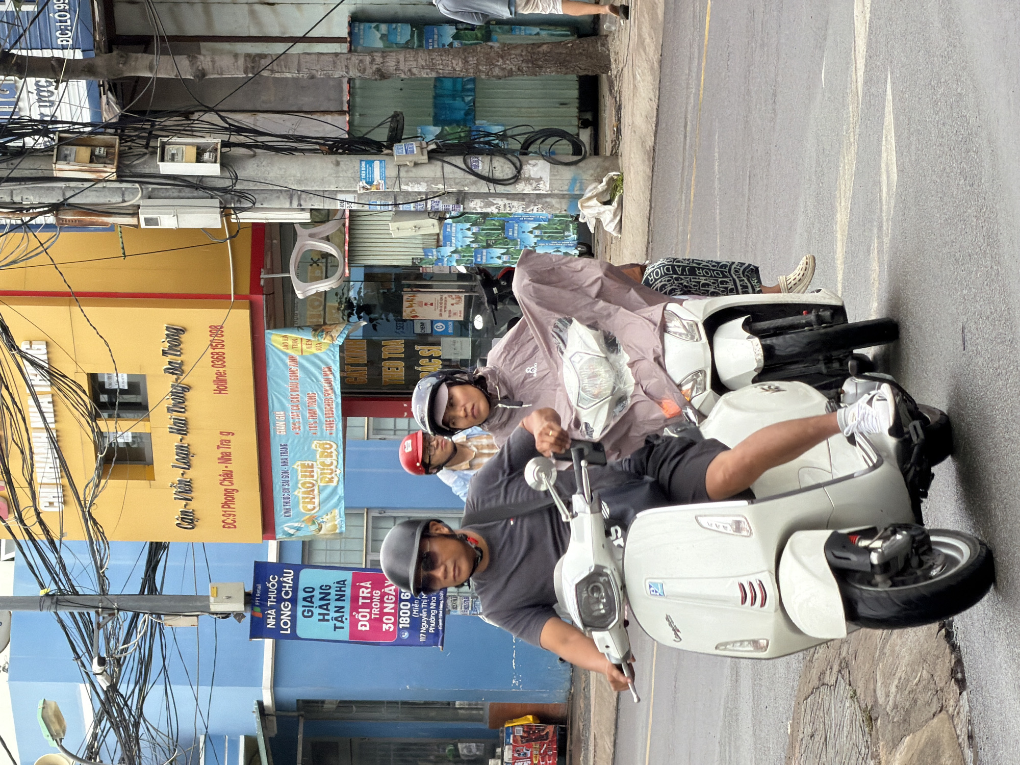 Two individuals are riding a white Vespa scooter on a wet city street in Nha Trang, Vietnam. The rider in front, a male, wears a black helmet, sunglasses, and a dark t-shirt with black shorts. The person behind him, likely female, wears a light purple rain poncho and patterned leggings. The scooter is white with chrome accents and has its headlights on.
The background features a blue building on the left and a yellow building on the right with signs in Vietnamese, indicating a commercial area. Electrical wires are prominently strung overhead, and a utility pole with electrical boxes is visible on the right. The street appears to be asphalt and shows signs of recent rain, with puddles and reflections. Other vehicles and pedestrians are not clearly visible in the immediate vicinity. The overall scene depicts a typical urban street environment in Vietnam.