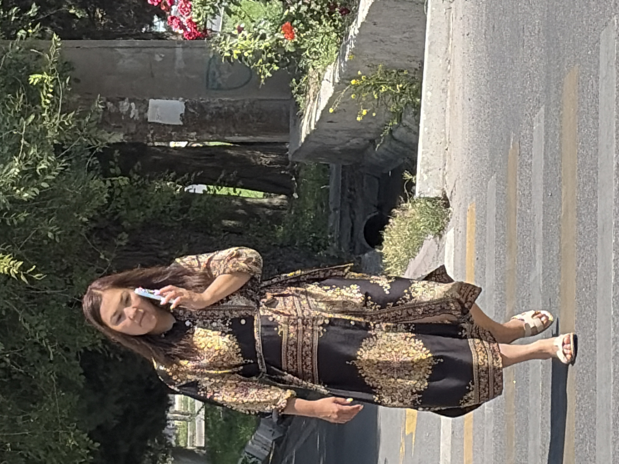 An adult female is walking across a street at a pedestrian crossing. She is wearing a long, patterned dress and sandals. She is holding a cell phone to her ear, engaged in a phone call. The background shows trees, a concrete wall, and some red flowers. The lighting suggests it is daytime with sunlight casting shadows. The street appears to be in an urban or suburban environment.