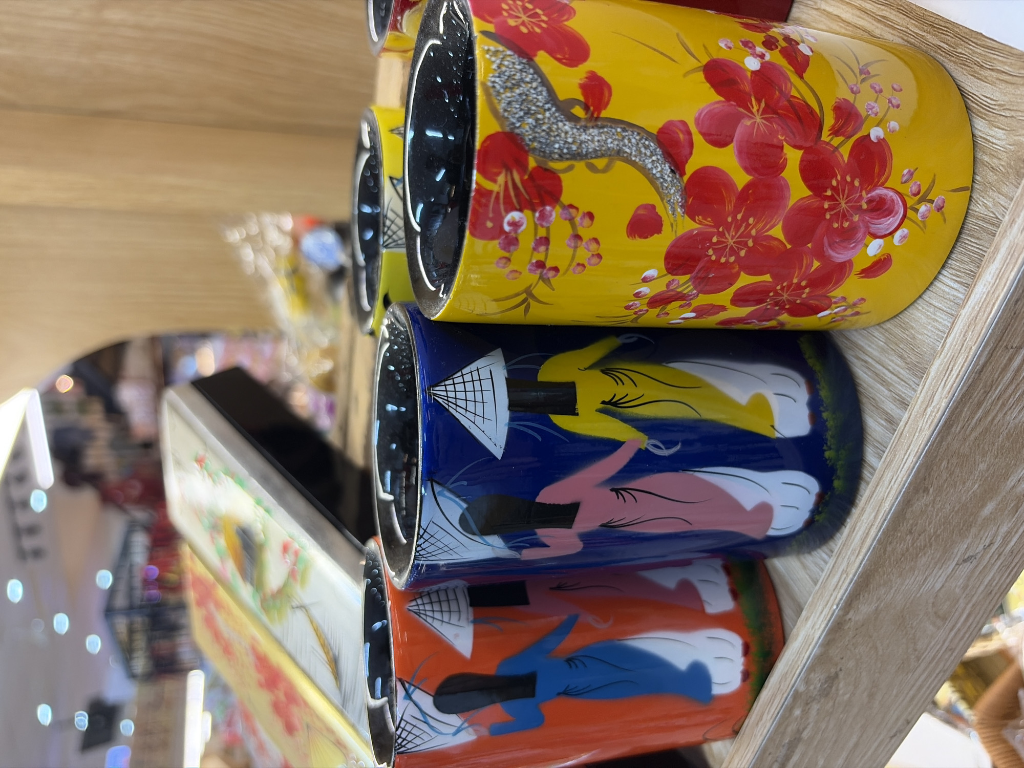 The image displays a close-up of several cylindrical ceramic cups arranged on a wooden shelf. The cups are decorated with colorful, hand-painted designs. The cup in the foreground is bright yellow with red floral patterns and a sparkling silver embellishment. To its left, a blue cup features a depiction of a person in traditional Vietnamese attire wearing a conical hat. An orange cup, to the left of the blue one, has a similar design with a figure in a blue dress and a conical hat. The background is blurred but suggests an indoor setting, possibly a shop or market, with shelving and other items visible. The lighting is bright, creating reflections on the glossy surfaces of the cups. The overall impression is of decorative souvenir items. The scene does not provide specific visual cues to determine its exact location within the city of Đà Nẵng, Vietnam, but the style of the artwork on the cups is consistent with Vietnamese folk art.