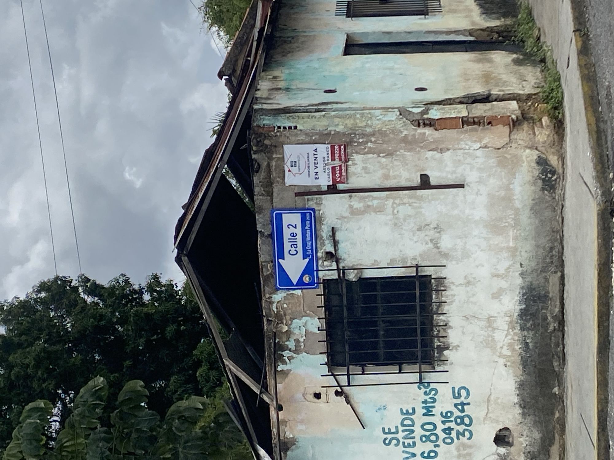 The image shows a run-down building in Tinaquillo, Venezuela. The building has a sign for Calle 2 and a sign that says "Se Vende" ("For Sale"). There is also a sign advertising a real estate agent. The building appears to be in a state of disrepair.