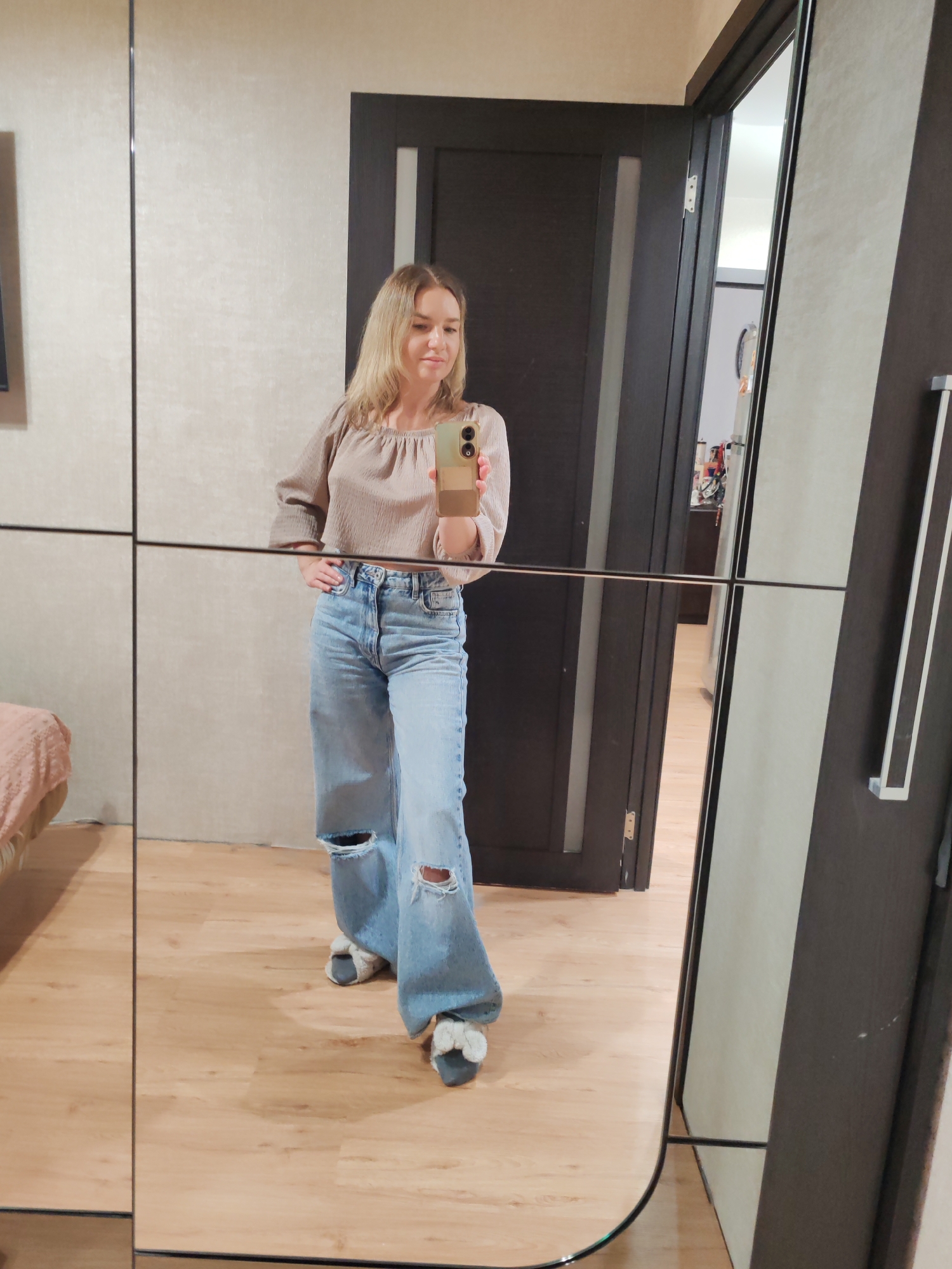 A full-length mirror reflection captures a woman standing indoors. She is fair-skinned with blonde hair, wearing a light brown, loose-fitting top and distressed, light blue jeans. She is holding a smartphone in her right hand, seemingly taking a selfie. Her left hand rests on her hip. She is wearing slippers.
Behind her, visible through an open doorway, is a glimpse of another room with furniture and other objects. The wall behind her is a neutral beige color.
Given the interior setting and the focus on personal appearance, there is no explicit evidence in the image to suggest a specific location within Petropavlovsk-Kamchatsky, Russia.