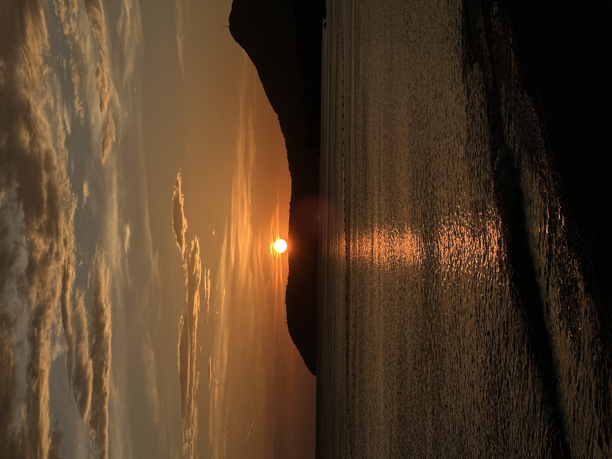 The image depicts a sunset over the Adriatic Sea in Bar, Montenegro. The sun, a bright orange orb, is partially obscured by a dark, silhouetted mountain range stretching across the horizon. The sky shows a gradient of orange hues, with long, striated clouds illuminated by the setting sun. The sea's surface reflects the sunlight, creating a shimmering, textured pattern of light. The right side of the image is considerably darker, suggesting the transition from daylight to twilight.
