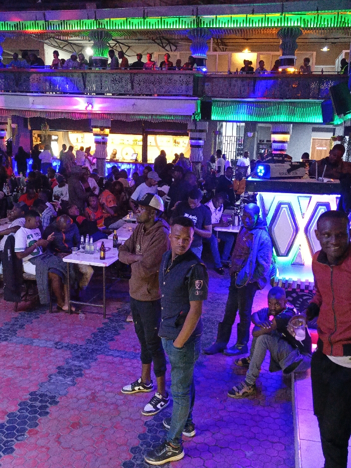 An indoor space houses a large crowd, possibly a club or bar, with a mezzanine level where people are visible. Tables with drinks are scattered throughout the main floor where most of the people are concentrated. A DJ booth glows with bright white and blue light. In the foreground, several men are standing and sitting. The space is illuminated with colorful neon lights, primarily green, blue, and purple. The floor is paved with patterned tiles of red, blue and grey.