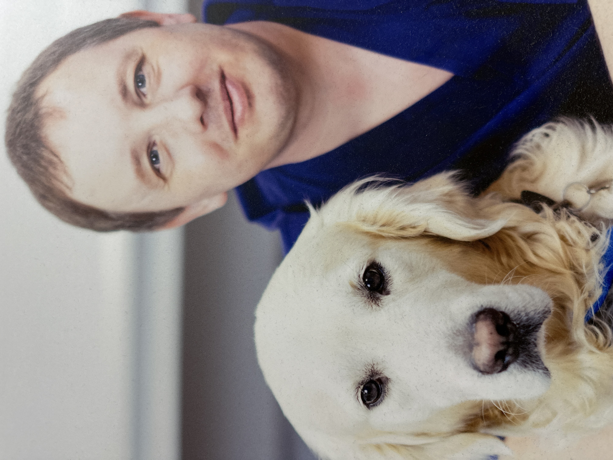 A man with short brown hair and blue eyes, wearing a blue uniform, is pictured with a golden retriever. The dog is positioned in front of the man, with its head and upper body visible. The man is holding the dog, and they appear to be posing for a photograph. The background is neutral, with a light gray wall and a hint of a darker surface below. The lighting is soft, creating a gentle illumination of the subjects. The image is a close-up portrait, focusing on the faces of the man and the dog. There is no indication of the city of Minsk, Belarus in the image.