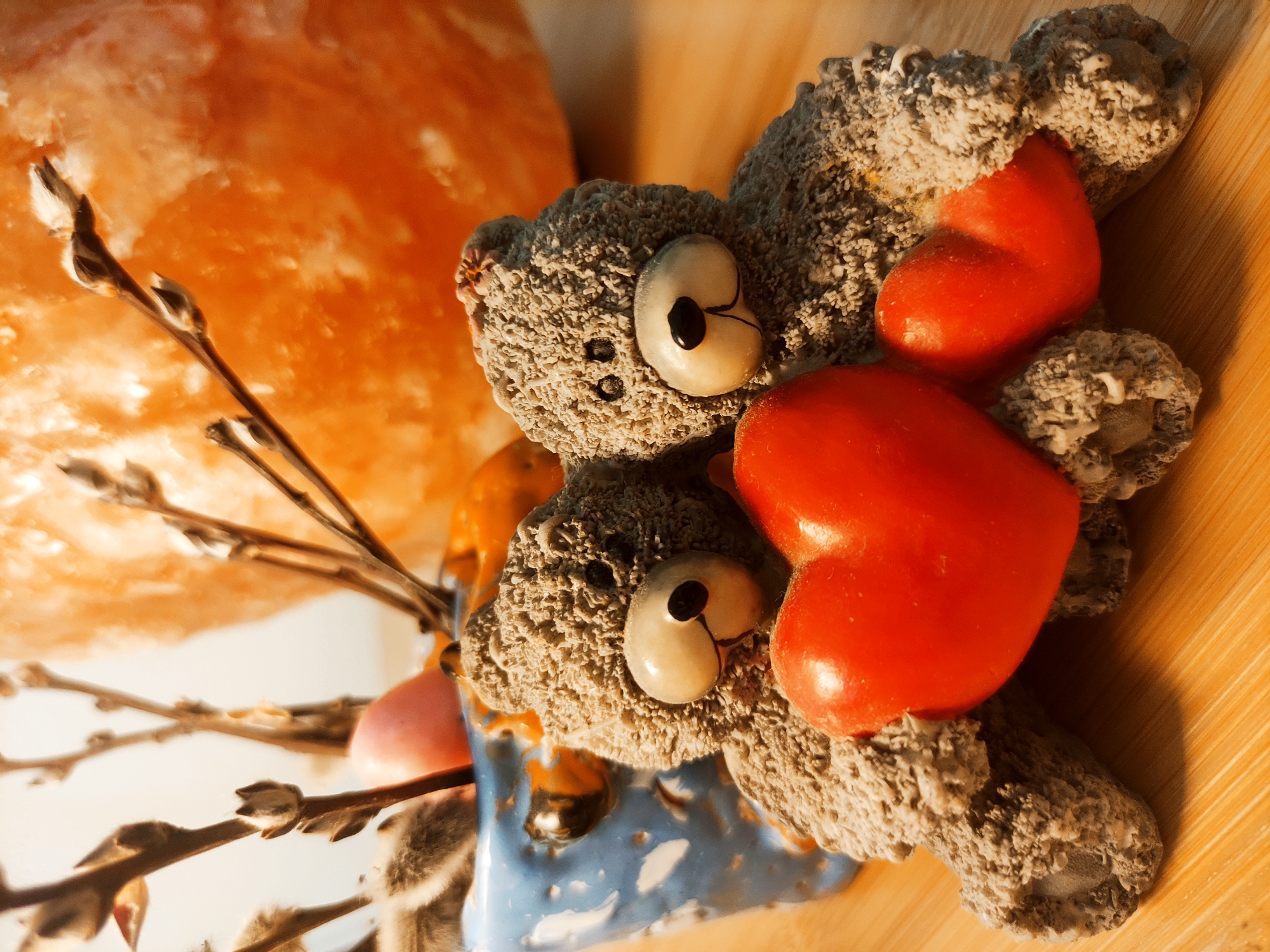Two gray teddy bears with painted eyes and two red hearts lie on a wooden surface. One bear is partially contained in a blue and white vase with catkins. A large orange mass, possibly a salt lamp, is visible in the background. The location is Unknown, Unknown.