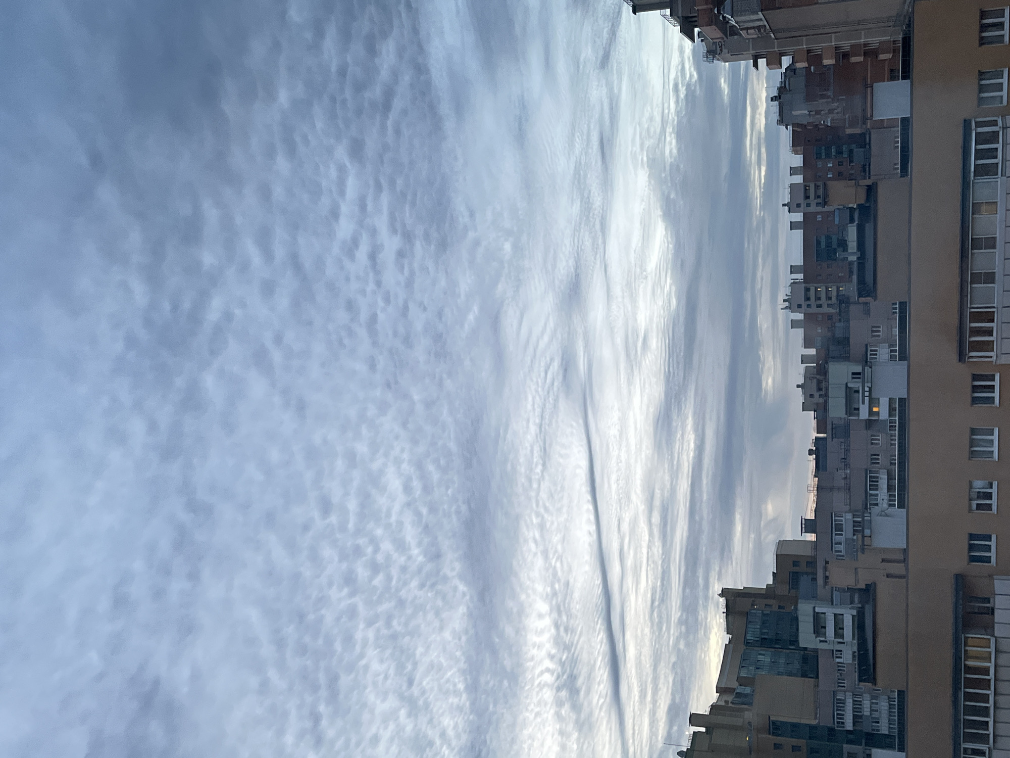 The image displays a cityscape with a cloudy sky. The buildings are of various heights and architectural styles, predominantly appearing to be apartment complexes or office buildings. The sky is filled with a textured pattern of clouds, ranging in color from light gray to dark blue, suggesting an overcast or dusk/dawn setting. The foreground features the tops of several buildings, some with visible windows and balconies. The overall scene depicts an urban environment under a dynamic sky.
