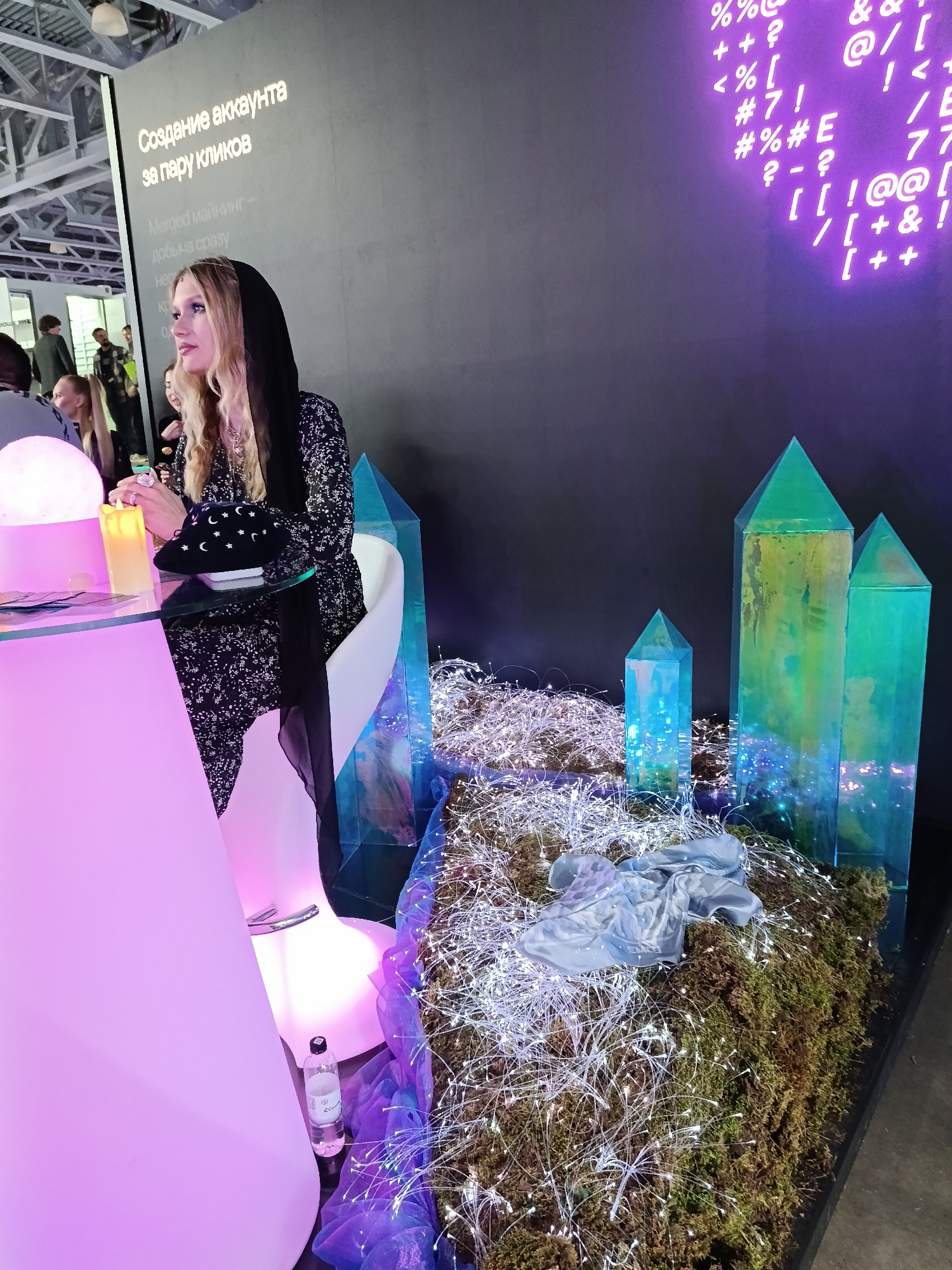 The image, taken in Khimki, Russia, shows a woman with long blonde hair sitting at a tall, pink-lit table. She is wearing a black and white patterned dress and a black hooded scarf. On the table, there is a black hat decorated with moons and stars, a lit artificial candle, and a glowing orb. A white chair is behind her, and a bottle of water is on the floor near the chair.
Behind the table, there is a black wall displaying white text in Cyrillic and neon purple symbols. To the right of the woman, a display features four tall, transparent, geometric shapes resembling crystals, arranged on a base of moss and adorned with fiber optic lights. A folded blue shirt rests on the display.