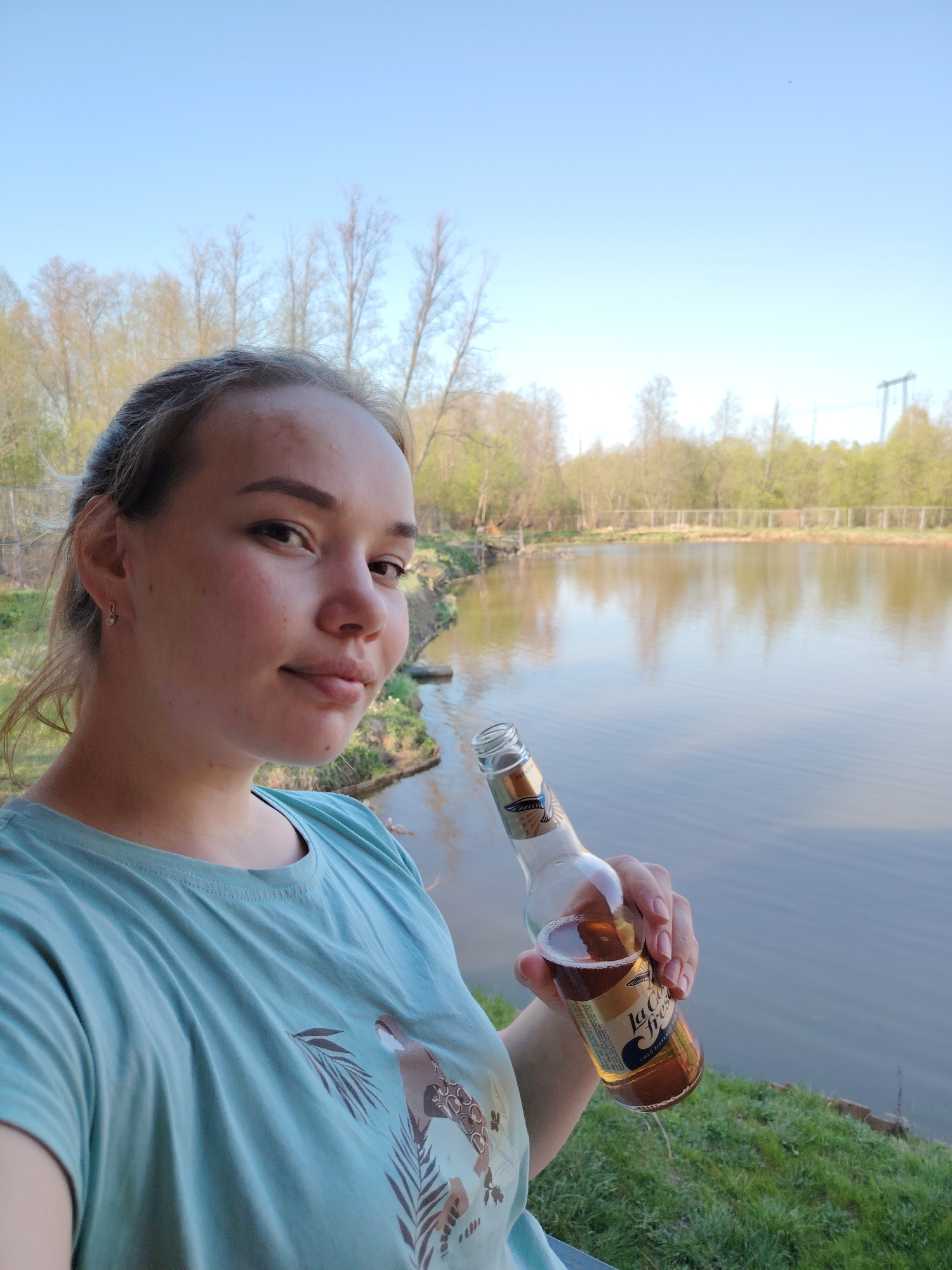 A woman is holding a beer bottle while standing near a lake or pond in Malyi Bukor, Russia. The woman is facing the camera with a slight smile. She has fair skin, light brown hair pulled back, and is wearing a turquoise t-shirt with a brown and black design. Her left ear is visible, revealing a small earring.
She is holding a glass beer bottle with a label that reads "la Co fresc". The bottle is partially empty, suggesting it has been consumed.
Behind her is a body of water reflecting the sky and surrounding trees. There is a grassy bank on the edge of the water, and trees are visible in the background. A wire fence or structure is partially visible on the far side of the lake, along with power lines. The sky is blue with some haze near the horizon, suggesting clear weather.