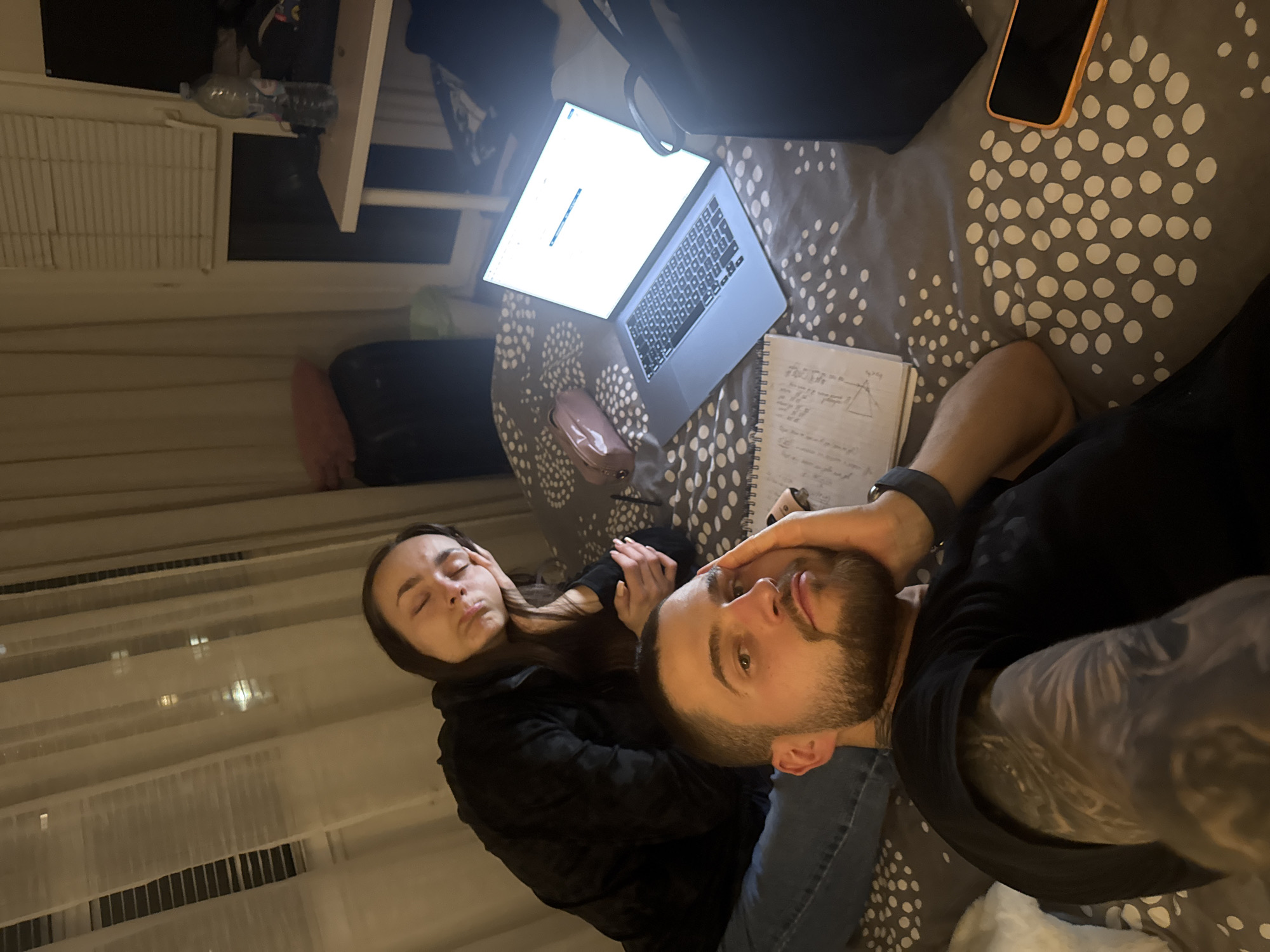 A couple is lying on a bed with a patterned grey and white duvet. A man with a beard and a tattooed arm is taking a selfie while holding the woman’s face. The woman has dark hair and appears tired. A laptop is open on the bed next to a notebook with handwritten notes and a brown pencil case. A phone with an orange case is also on the bed. In the background, there are curtains and a window. The location is unclear, and there is no indication this photo was taken in Sofia, Bulgaria.