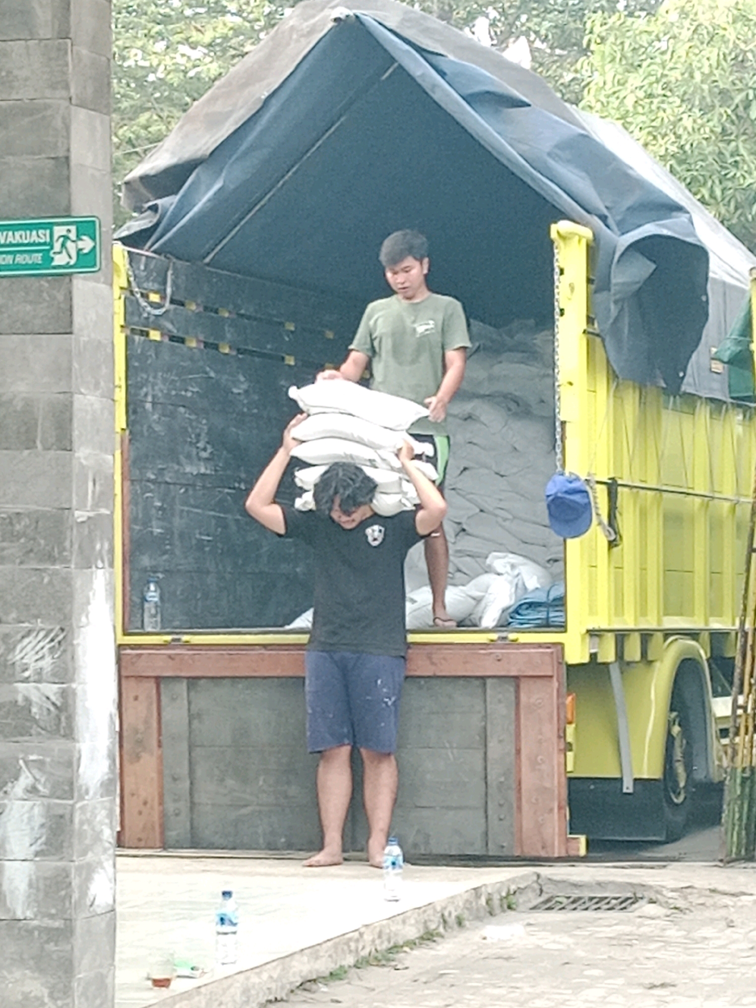 In Salatiga, Indonesia, two men are observed near the open rear of a yellow truck, which is covered with a dark tarp. One man, wearing a green t-shirt, stands inside the truck's cargo area, holding a stack of white sacks. Another man, in a black t-shirt and blue shorts, stands on a wooden plank at the rear of the truck, carrying a similar stack of white sacks on his shoulders. A blue cap hangs from a chain inside the truck. To the left of the truck, a grey brick wall features a green sign with white text and an evacuation symbol. The ground is paved, and several water bottles are visible on the ground in the foreground.