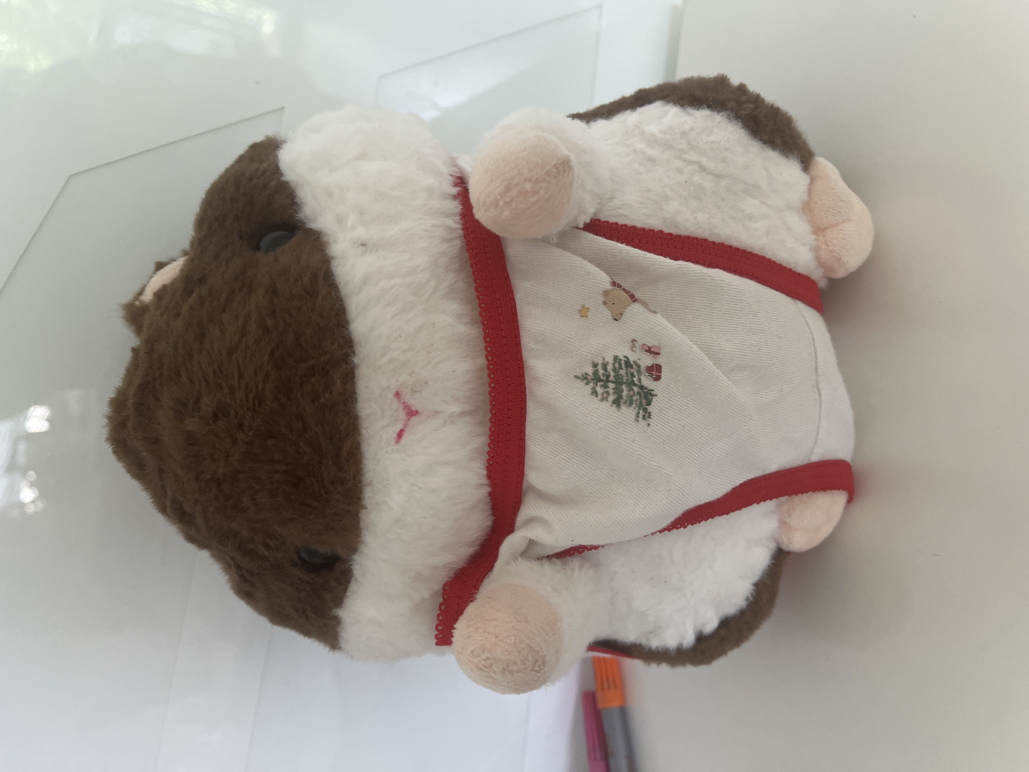 A brown and white plush toy, resembling a guinea pig, is depicted lying on a light-colored surface. The toy's head, which is predominantly brown with a black button-like eye, faces the left side of the frame. Its body is covered in patches of brown and fluffy white fur.
The plush toy is dressed in a festive, light-colored garment, possibly a shirt or a blanket, that features red scalloped trim along its edges. This garment is embroidered with Christmas-themed imagery. A green Christmas tree, topped with a yellow or red star, is prominently displayed on the lower part of the clothing. Above the tree, a small brown figure, resembling a bear or a reindeer cub wearing a red hat, is also embroidered, along with a smaller yellow star.
Small, rounded, light pink ears and feet are visible, peeking out from beneath the fur and the edges of the garment. The background is a soft, out-of-focus white, suggesting an indoor setting with possibly a wall or another surface. Reflections hint at a smooth, potentially glossy surface. In the bottom right corner of the main image, two markers, one orange and one purple, are partially visible on the surface. The plush toy is stationary, and no specific activity is taking place. There is no discernible text in the image.