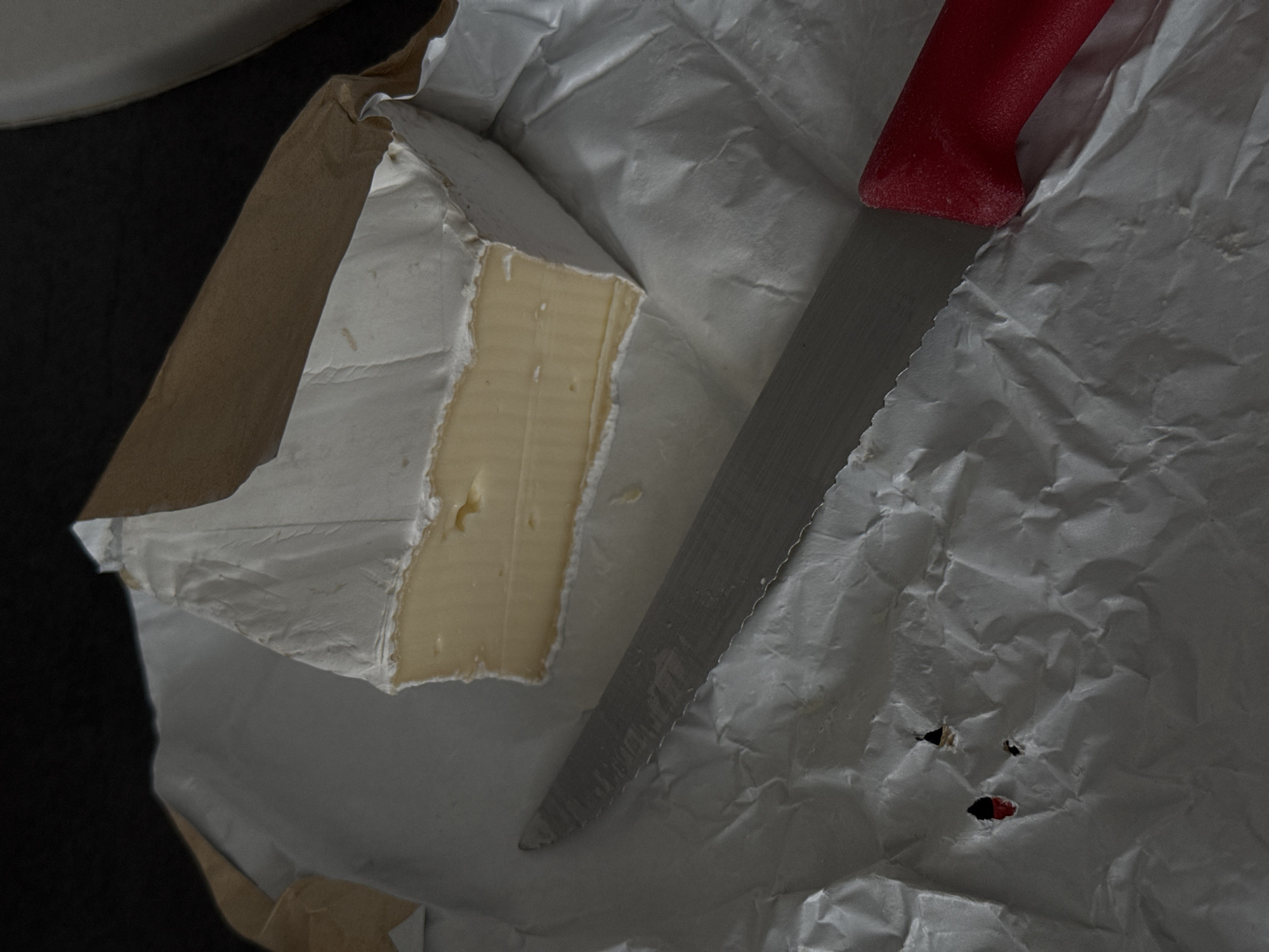 A close-up, overhead shot shows a wedge of brie cheese resting on crumpled white paper. The cheese has a white rind and a pale yellow interior. A serrated knife with a red handle lies diagonally across the paper in front of the cheese. The background is dark and out of focus. The scene is not in Siegburg, Germany.