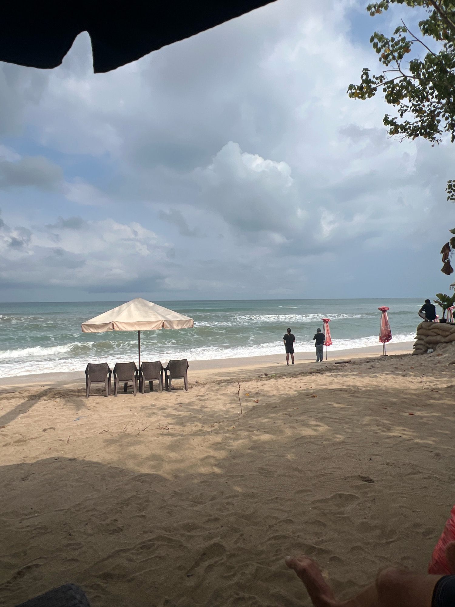 The image depicts a beach scene in Kuta, Indonesia, under a cloudy sky. In the foreground, a sandy beach is visible, with shadows cast by unseen objects. Four empty plastic chairs are arranged in a row under a large, beige beach umbrella, positioned near the water's edge. The ocean has moderate waves rolling onto the shore. Further down the beach, two figures stand near the water, facing the sea. Two red beach umbrellas are also visible. On the right side of the image, a person is seated on sandbags, and some greenery is present. The overall atmosphere suggests a quiet, possibly overcast day at the beach.