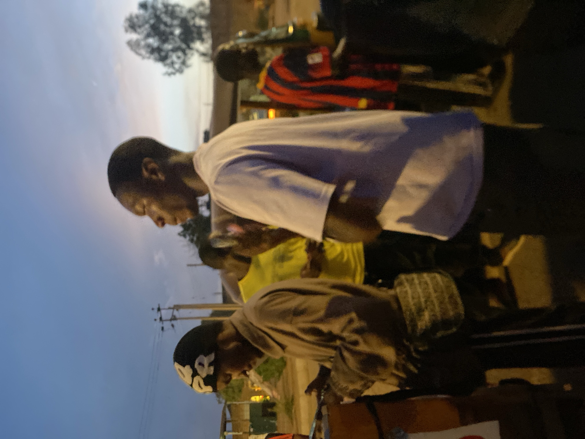 This image captures a street scene at twilight in Bukuru, Nigeria, showing five male figures under a gradient sky transitioning from blue to a lighter hue.
In the foreground, on the left, a male figure wearing a dark beanie with white "PR" lettering, a light brown long-sleeved shirt, and dark pants with vertical white stripes is partially visible. He has a patterned cross-body bag and is looking downwards, with his right hand extended towards an unseen object or surface. Immediately in front of him is a low, reddish-brown structure, possibly a counter or stall.
A central male figure stands in profile, facing left, wearing a plain white short-sleeved t-shirt and dark trousers. His arms are bent at the elbows. He has a solemn expression.
Behind the central figure, slightly to the left, another male figure is partially visible, wearing a bright yellow t-shirt. Further to the right in the background, a male figure wearing a red and black striped shirt stands near a yellow and green vehicle, possibly a commercial tricycle or tuk-tuk. Another male figure is dimly visible to the far right.
The background includes utility poles with wires against the sky and a distant tree with sparse foliage to the right. The ground is dimly lit, suggesting artificial light sources are active, casting warm yellow and orange tones. Distant structures are faintly visible under the fading light.