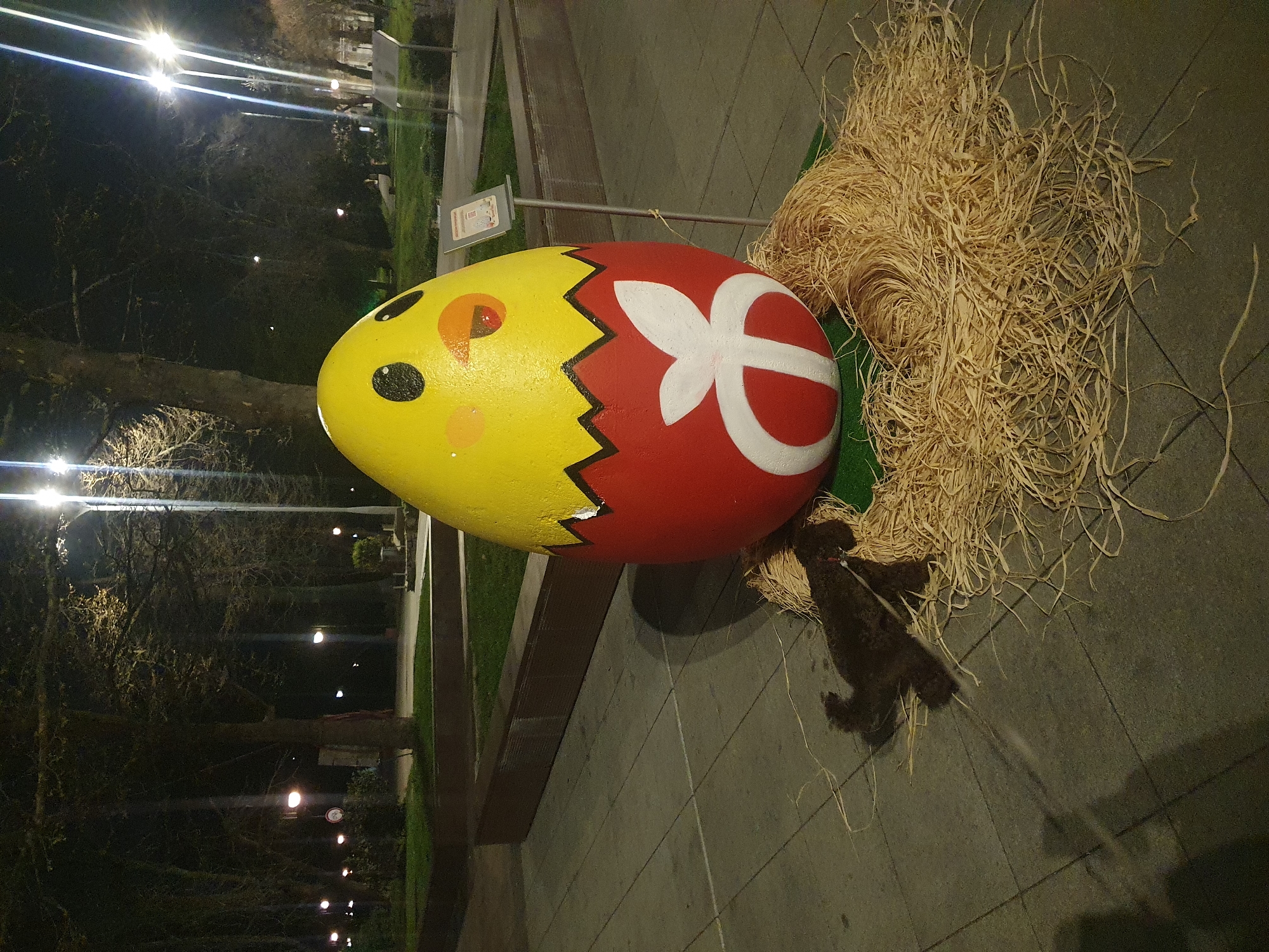 The image shows a large, painted Easter egg sculpture on display in a public space. The egg is divided into two sections: a yellow section with a chick's face (eye, beak, and cheek) and a red section with a white stylized flower design. The two sections are separated by a jagged black line. The sculpture is surrounded by a pile of straw-like material. A small, dark-colored dog is near the sculpture. The sculpture appears to be on a sidewalk or paved area. Trees are visible in the background, along with streetlights suggesting it is night or dusk. This is probably located in the city of Plovdiv, Bulgaria, as per the user’s instruction.
