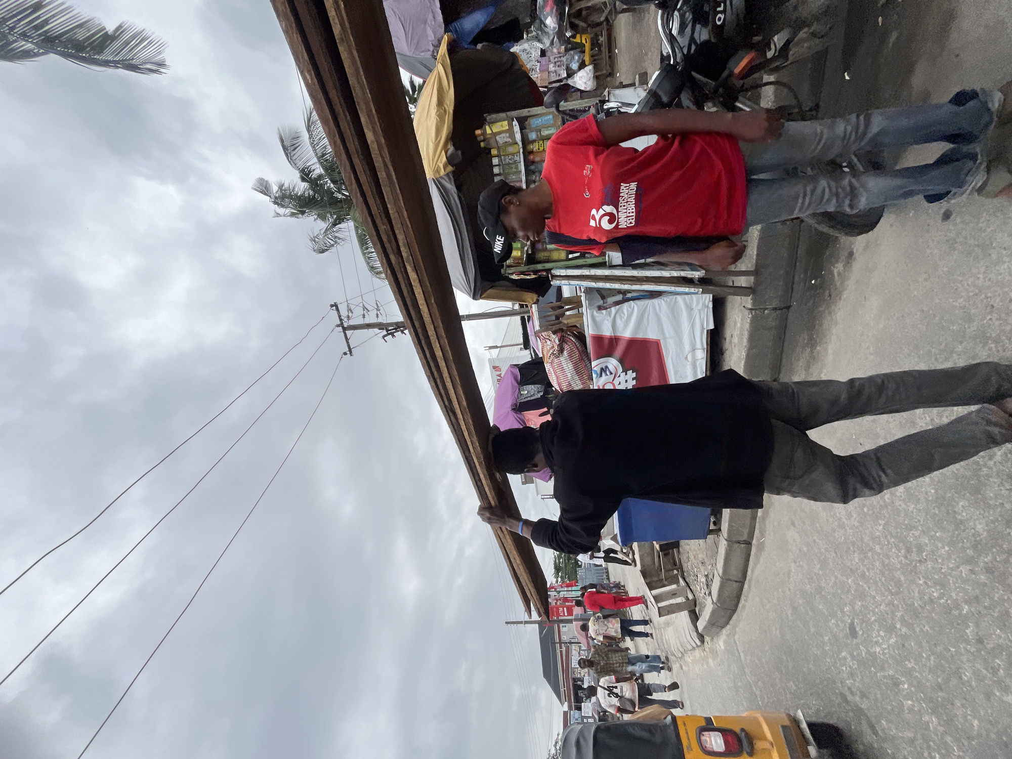 A man in a black shirt and grey pants carries a long wooden plank on his head while walking on a paved street. To his right, a woman in a red t-shirt and blue jeans stands near a market stall. In the background, other pedestrians walk along the street. The scene takes place in a busy, outdoor market in Ado, Nigeria, with various stalls, vehicles, and utility poles visible. The sky is overcast.