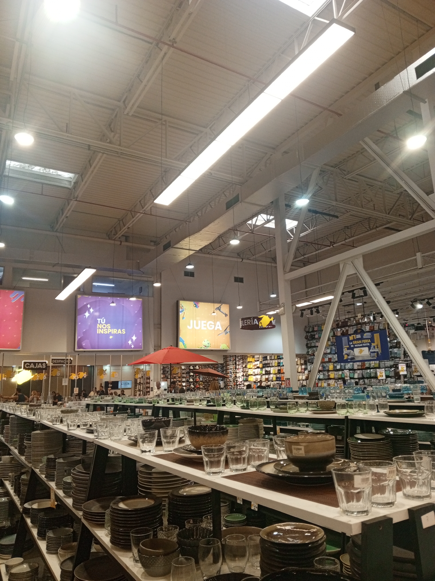 A large retail store in Pampatar, Venezuela, presents shelves stocked with dinnerware, including stacks of plates, bowls, and glassware. Signs with phrases like "Tú nos inspiras" and "Juega" are visible. Several people are browsing and shopping in the background. The interior features high ceilings with exposed metal beams and fluorescent lighting. An orange umbrella is also visible in the store.