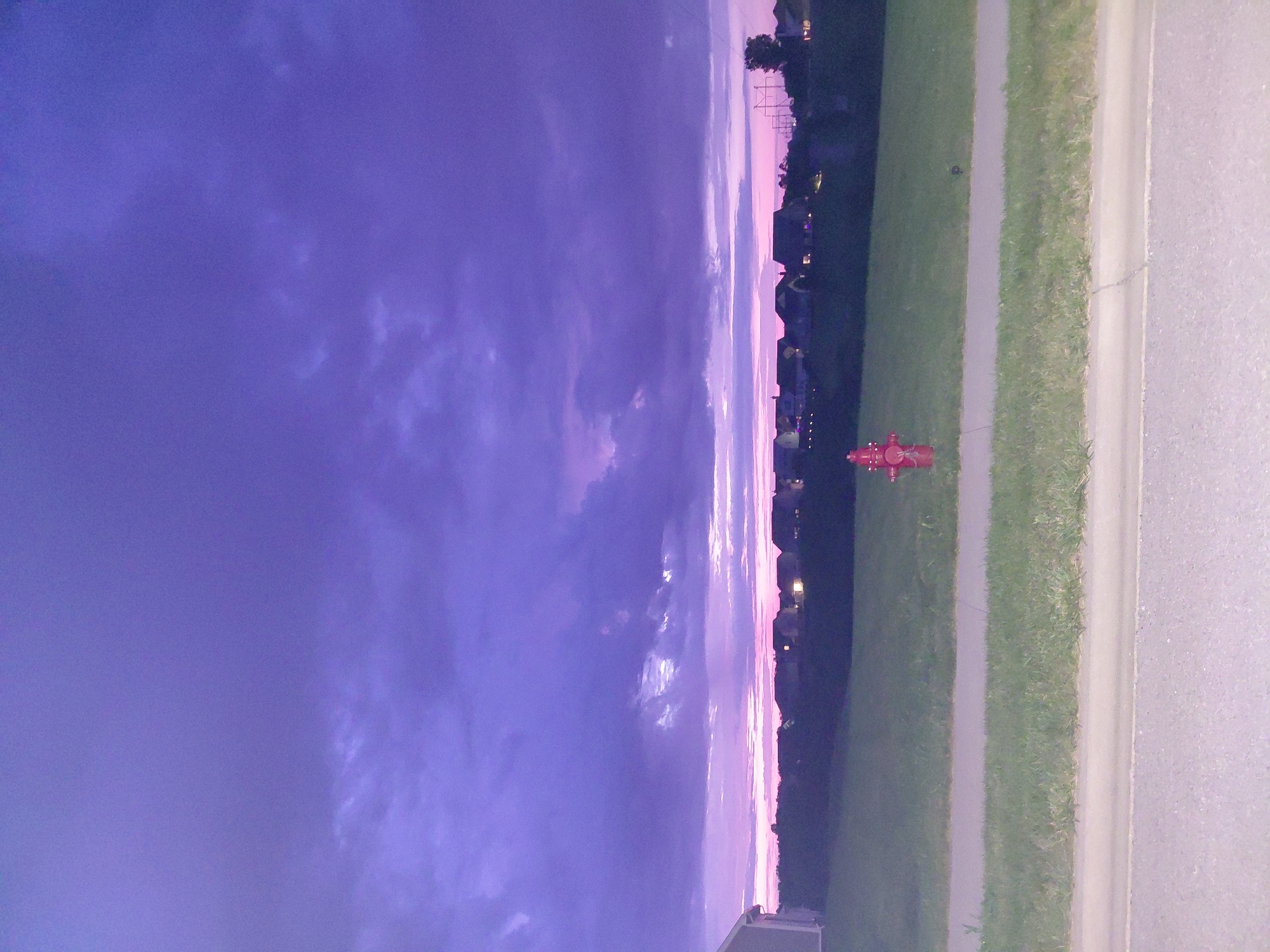 The image depicts a suburban residential area at dusk, illuminated by a vibrant purple and pink sky. A row of houses with illuminated windows stretches across the horizon. In the foreground, a fire hydrant stands in a grassy field. A paved road and sidewalk are visible in the lower portion of the image. The scene appears peaceful and quiet. The location is stated to be in the city of Richmond, United States of America.