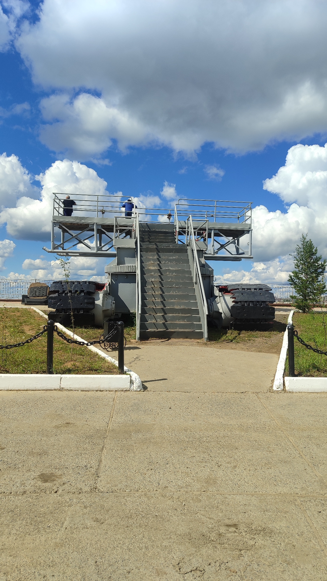 The image depicts a monument in the city of Mirny, Russia, consisting of a large metal structure with stairs leading to an elevated platform. Two individuals, one wearing a dark shirt and the other in a blue shirt, are standing on the platform. The structure appears to be an old piece of industrial equipment, possibly related to mining, as evidenced by the large, dark caterpillar tracks visible on either side of the base. The monument is situated outdoors, with a paved walkway in the foreground, a grassy area with a chain fence, and a small evergreen tree to the right. The background features a bright blue sky with fluffy white clouds. The overall scene is one of historical preservation or a public display of industrial heritage.