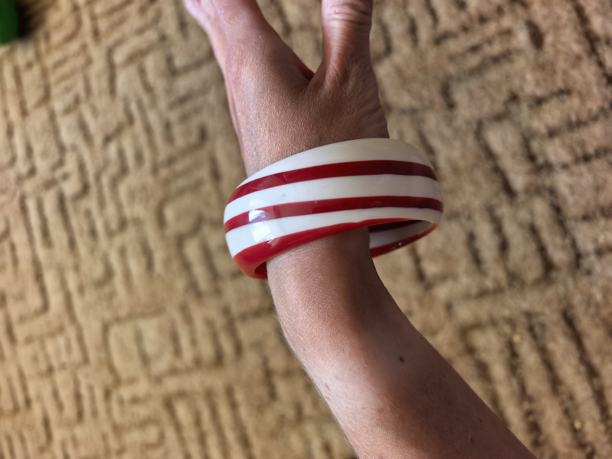 A close-up shot focuses on a person's forearm and hand adorned with a thick, striped bracelet. The bracelet alternates between white and red bands. The person's hand is partially visible, with fingers slightly bent. The background is a blurred, textured surface, possibly a carpet or fabric with a repetitive pattern. There is no evidence to suggest the image was taken in Sterlitamak, Russia.
