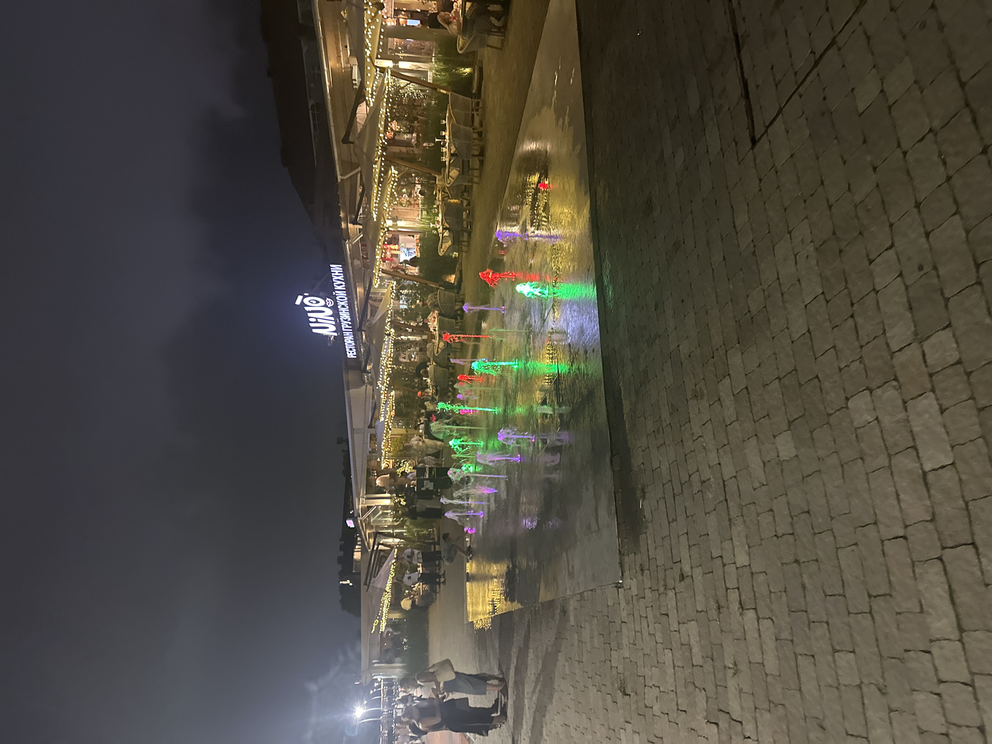 The image shows an outdoor dining area at night. A man is bent over a colorful illuminated fountain in the foreground. In the background, there is a restaurant with outdoor seating. The restaurant has a neon sign that says "NINO" and text in Russian that appears to be a restaurant name. People are seated at tables under umbrellas. The ground is paved with cobblestones. The city of Sirius, Russia is known for its modern architecture and public spaces, often featuring illuminated fountains and vibrant nightlife.