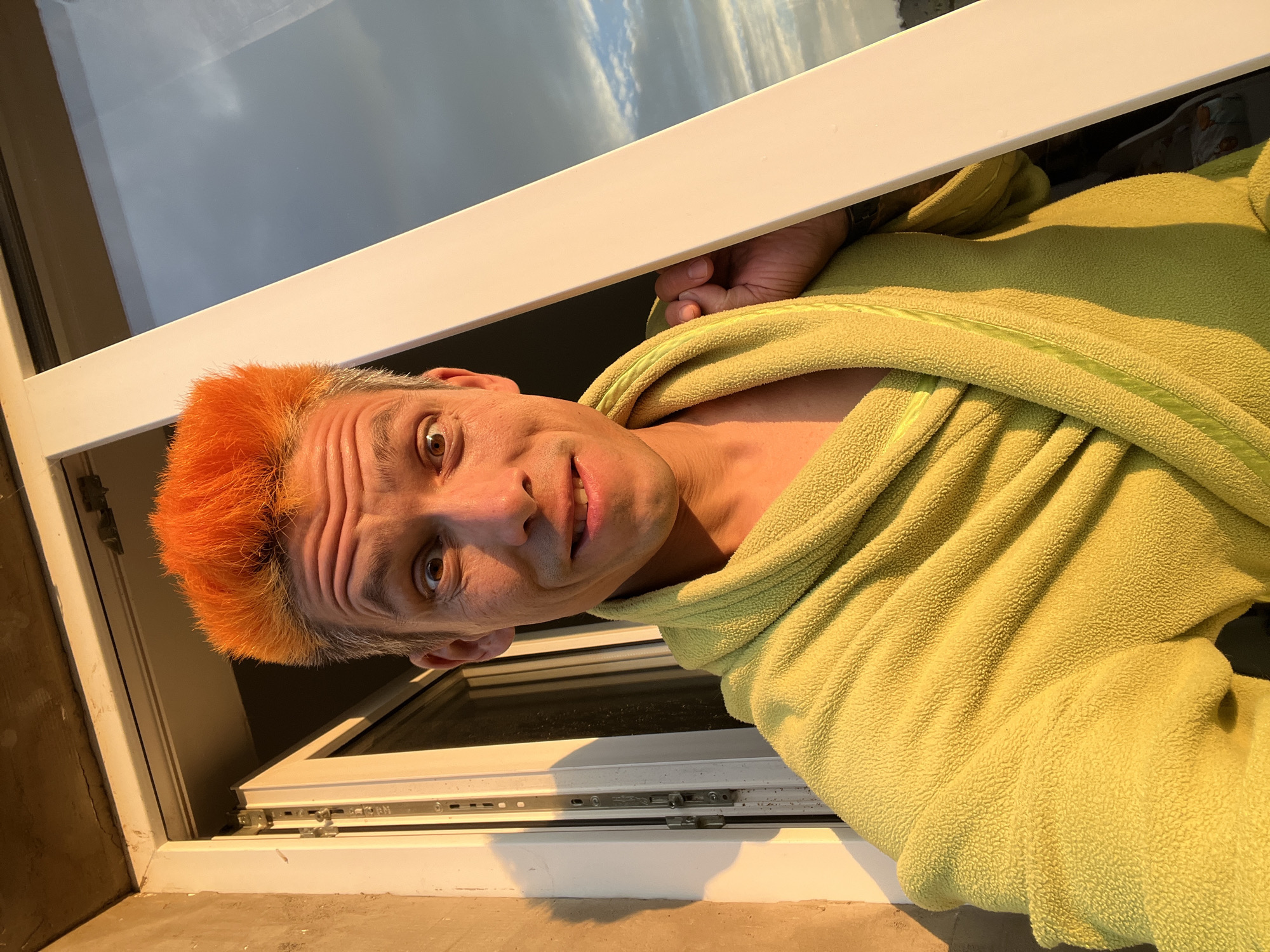 The image is a selfie taken by a man with bright orange hair, wearing a lime green bathrobe. He is standing in front of an open window, with the warm glow of sunset illuminating his face and the left side of the frame. The man is making a surprised or playful expression, with wide eyes and a slightly open mouth. A white window frame bisects the image vertically on the right side. Through the window, a cloudy sky is visible, reflecting the golden hour light. The wall on the left appears to be made of rough concrete. The overall impression is that of a candid moment captured during a beautiful sunset, possibly at a residence in Chaikovsky, Russia, given the context provided.