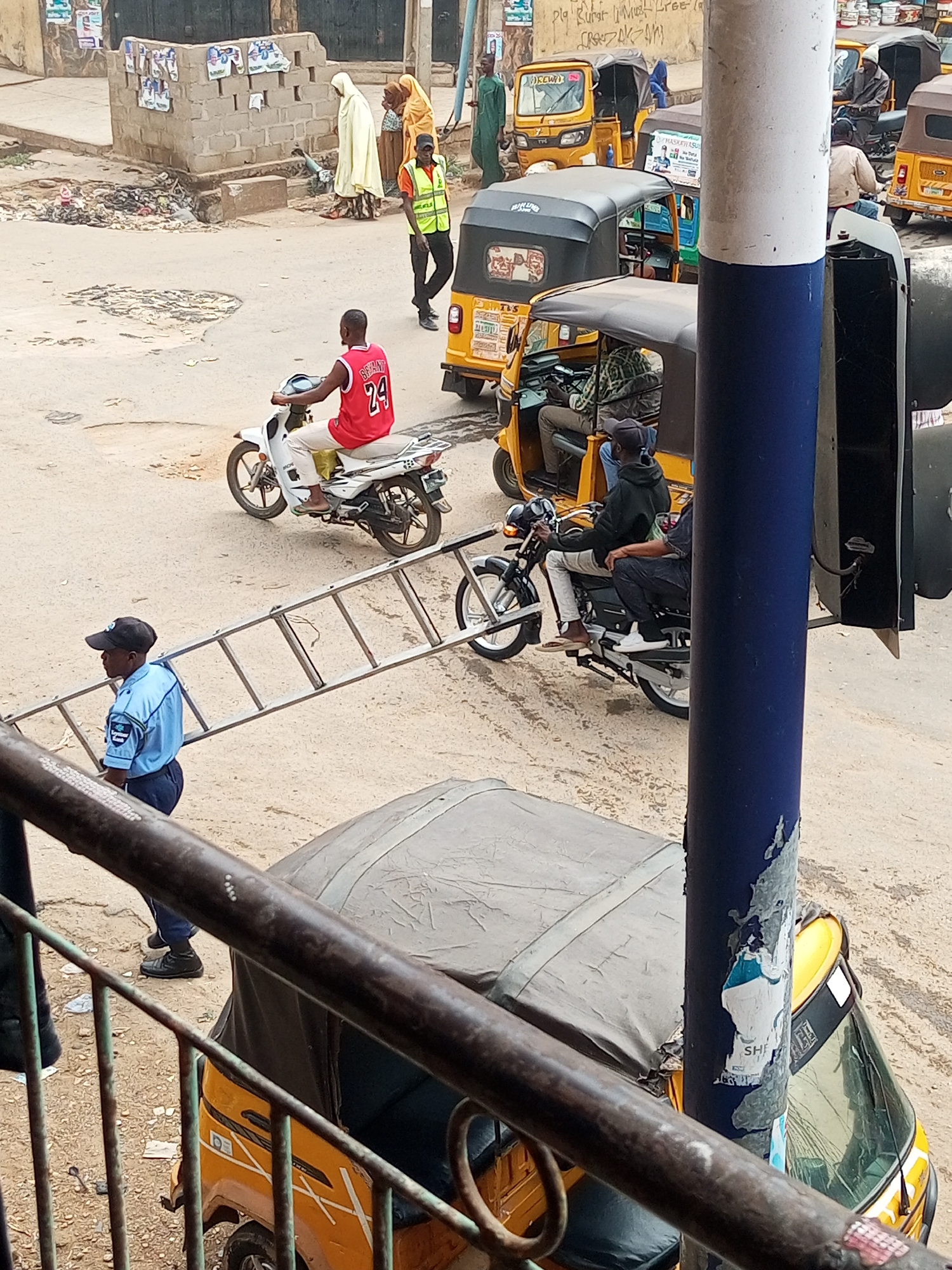 The image depicts a street scene in what appears to be the city of Maiburiji, Nigeria. In the foreground, there is a partially obscured view through a metal railing. Behind the railing, a yellow three-wheeled taxi (keke) is visible. The middle ground shows a busy street with several other kekes, motorcycles, and pedestrians. A man in a blue uniform is carrying a ladder horizontally across the street. A man on a white motorcycle is wearing a red tank top with the number 24. In the background, there are buildings and more pedestrians, some wearing traditional Muslim garments. The street is unpaved and dusty, with some litter visible.