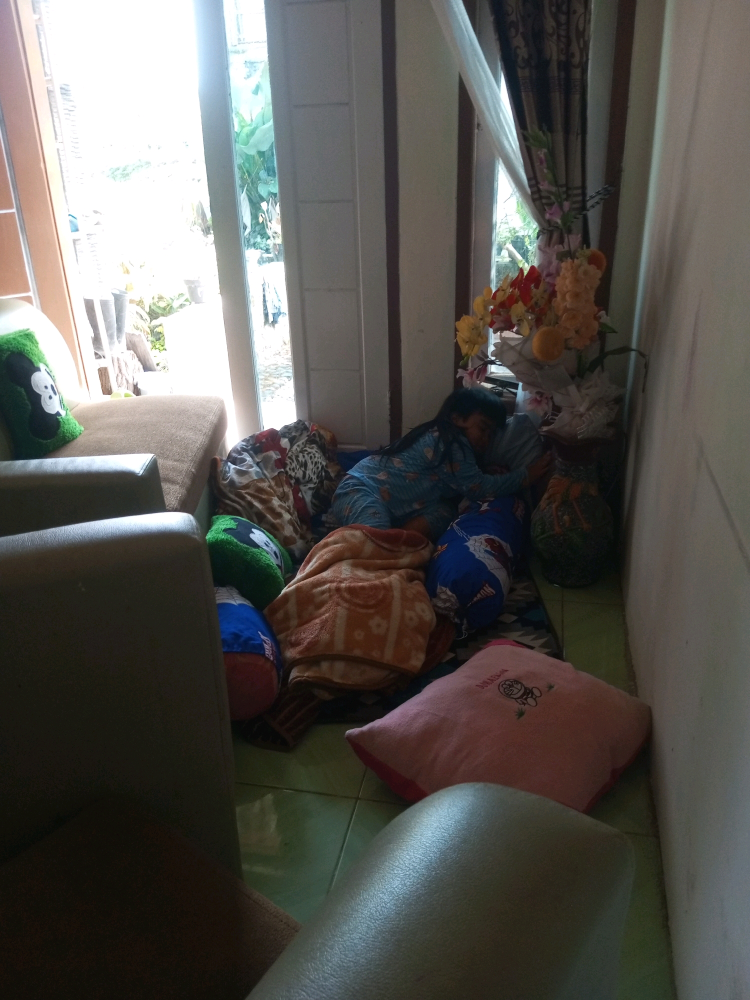 A young child, wearing blue pajamas, sleeps on the floor in a room in Krasak, Indonesia, surrounded by blankets and pillows. The child is nestled on a patterned mat, with a blue pillow patterned with what appears to be cartoon characters. To the child's left, a pile of blankets includes one with a cheetah print and another with a floral pattern. A green pillow with a black and white cartoon character is visible to the left, near a tan couch. A large pink pillow with a cartoon character embroidered on it is on the floor in the foreground. The room features a door and a window, with light streaming in from the outside. A vase with artificial flowers sits on a dark surface near the child. The walls are painted a light color, and the floor appears to be tiled.
