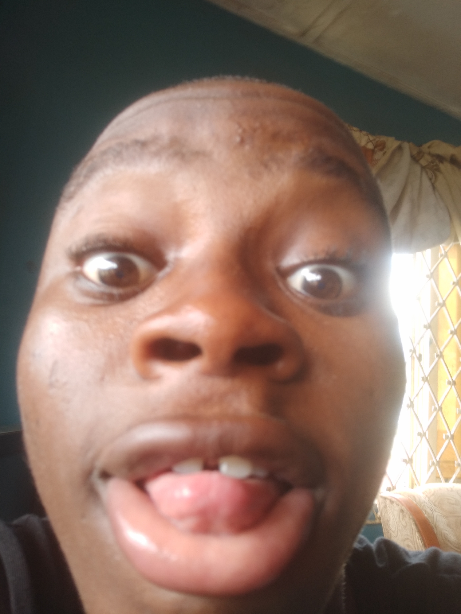 A close-up self-portrait of a person with dark skin. The person has a shaved head, wide eyes, and is sticking their tongue out. Their mouth is open and lips are glossy. The background is partly obscured, showing a teal wall and a window with patterned curtains. The location is inferred to be in Epe, Nigeria. No specific details within the image confirm that Epe, Nigeria is the true location.