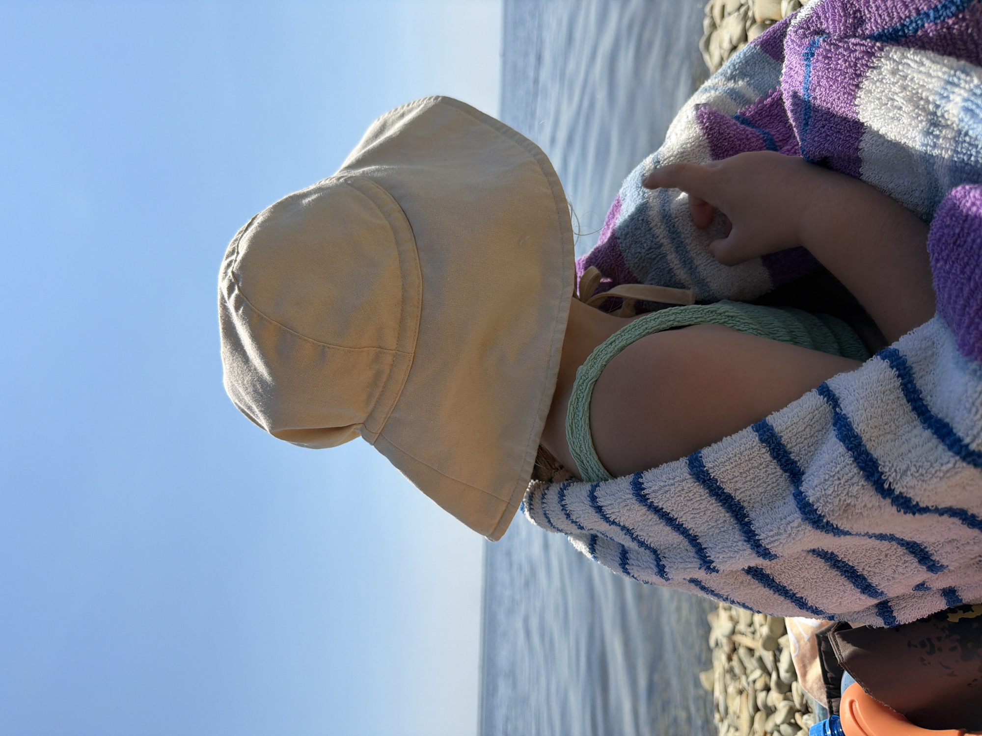 A person seen from behind, wearing a light-colored wide-brimmed hat and a light green top, is wrapped in a striped towel. They are sitting on a pebble beach with a body of water and a clear blue sky in the background. The scene is likely a beach on the Volga River in Kamyshin, Russia.