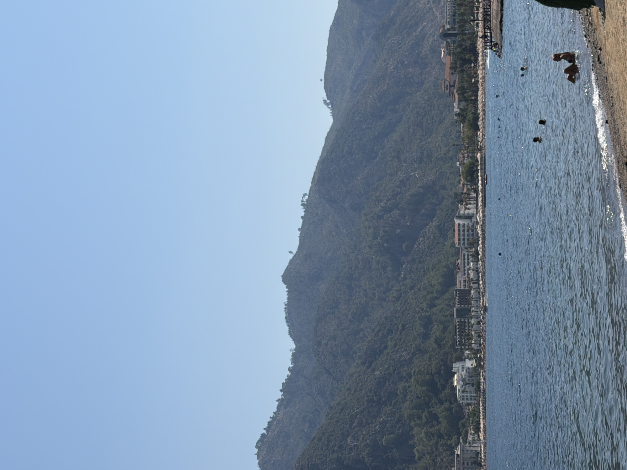 The image shows a view of the Marmaris coastline in Turkey. The foreground features a sandy beach and the blue waters of the sea. Several people are visible in the water, some swimming and others relaxing. In the middle ground, a line of hotels and resorts stretches along the coast, with their white facades and red-tiled roofs standing out against the lush green mountains in the background. The mountains themselves are covered in trees and vegetation. The sky is clear and blue, indicating a sunny day. A pier or jetty is visible on the right side of the image, with people on it. The overall scene depicts a popular tourist destination with a blend of natural beauty and man-made development.