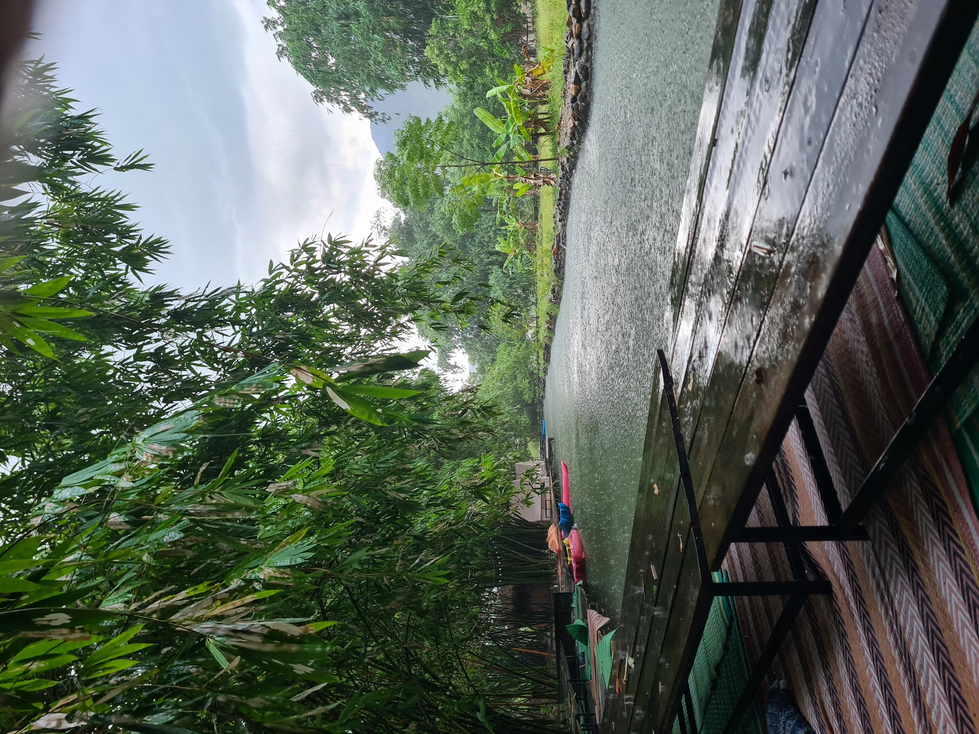 The image depicts a serene outdoor scene, likely a resort or a quiet riverside spot, on a rainy day in Da Nang, Vietnam. In the foreground, a wet wooden table and benches are placed on woven mats. The table surface is glistening with raindrops. Beyond the table, a calm body of water stretches towards the horizon. Several colorful kayaks are docked along the water's edge, indicating a recreational area. The left side of the scene is dominated by lush green bamboo foliage, with some leaves showing signs of browning. In the background, a verdant landscape unfolds, featuring a variety of trees, including banana plants, and a distant mountain shrouded in mist. The sky is overcast and grey, typical of rainy weather. The overall atmosphere is tranquil and natural, enhanced by the rain.